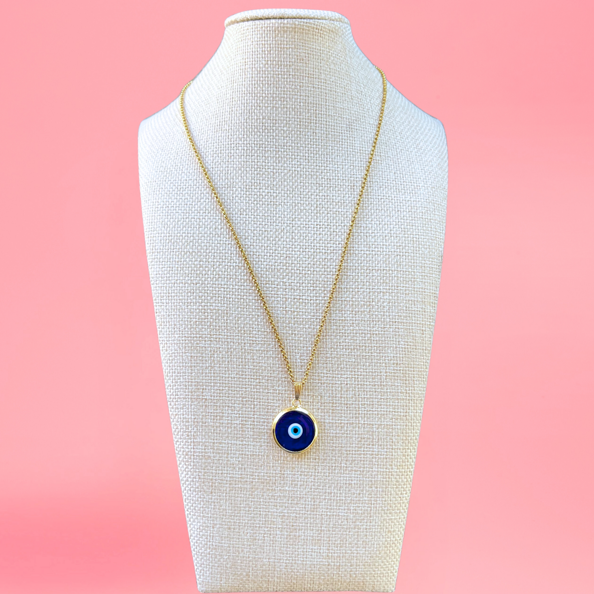 dark blue handmade turkish evil eye necklace modeled on beige bust with pink background. lampwork glass evil eye framed in gold-plated stainless steal with gold-plated stainless steel chain. view from the front