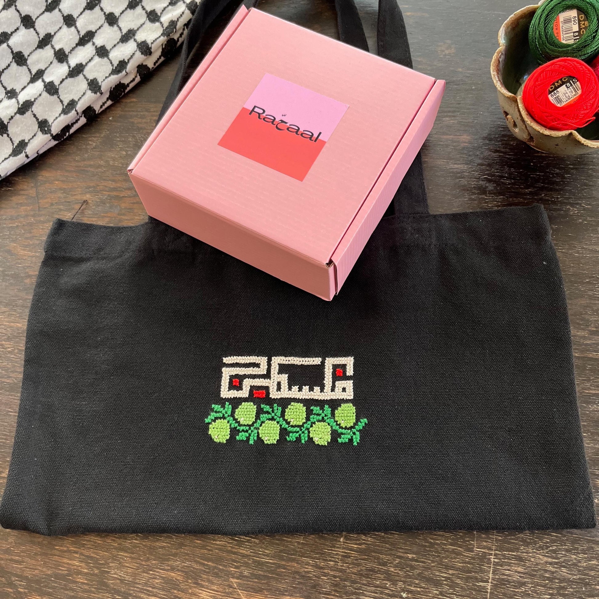 Tatreez tote bag kit with olive design. black tote bag, pink Rahaal tatreez kit box and embroidery threads, view from above