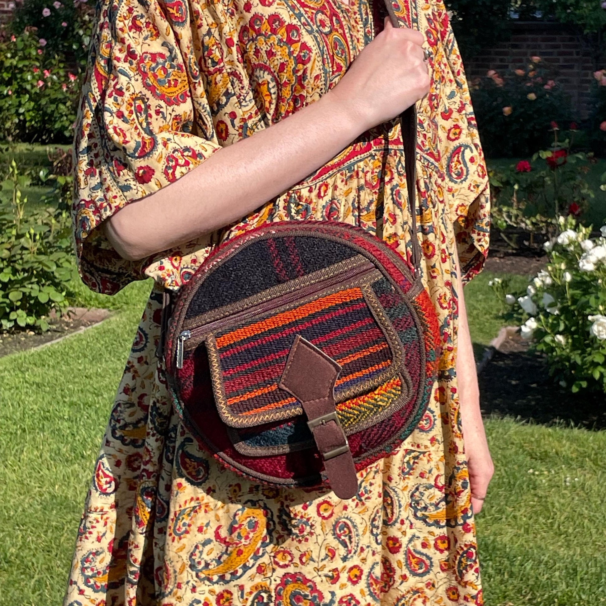 view from the front of a colorful circle bag modeled on a woman in a garden. the purse has handwoven sections of wool, trimmed with brown fabric. The bag has a long strap, front pockets, zippers, and a faux buckle over a snap closure