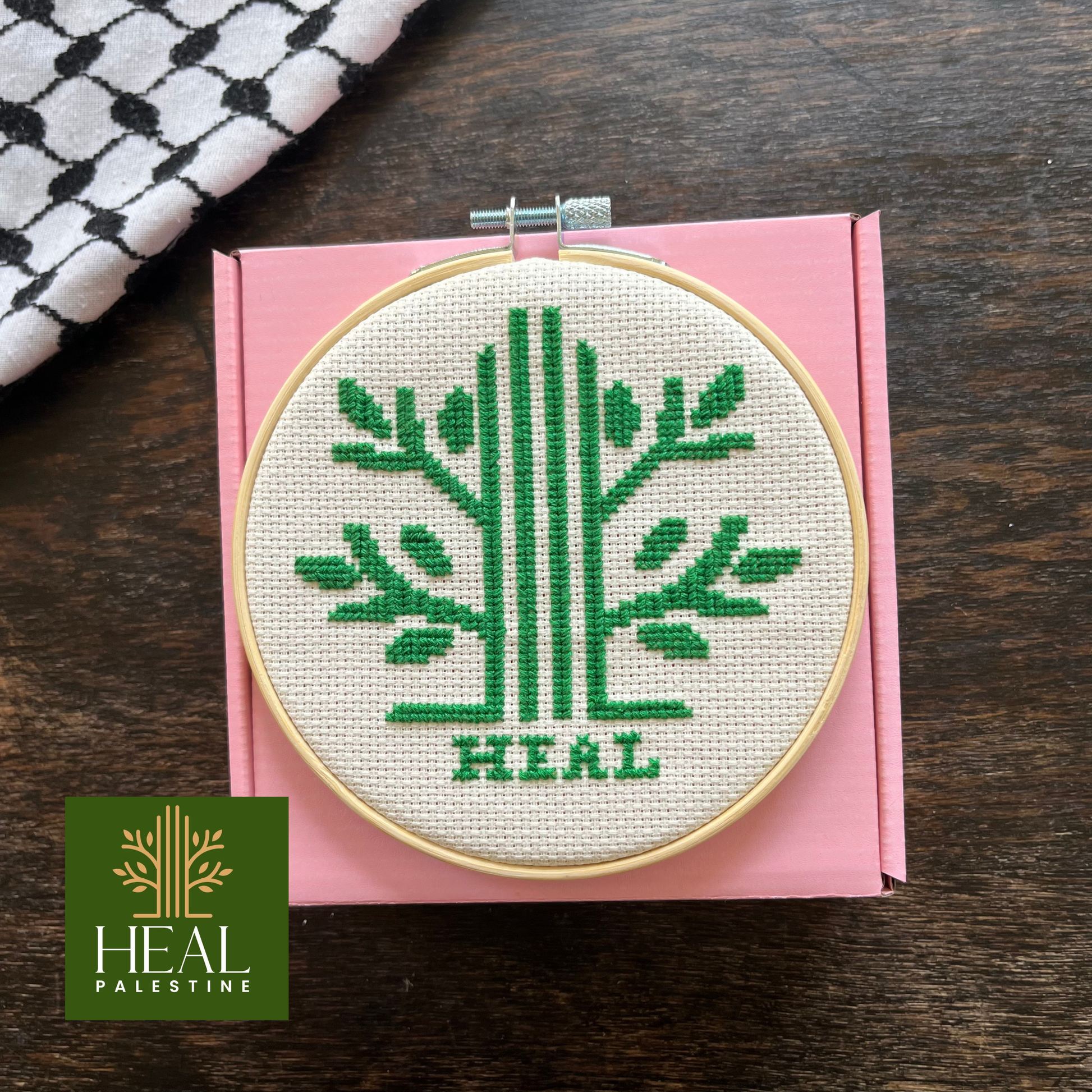 Tatreez kit with Palestinian olive tree design. white aida cloth, embroidery hoop, pink Rahaal tatreez kit box and embroidery threads, view from above