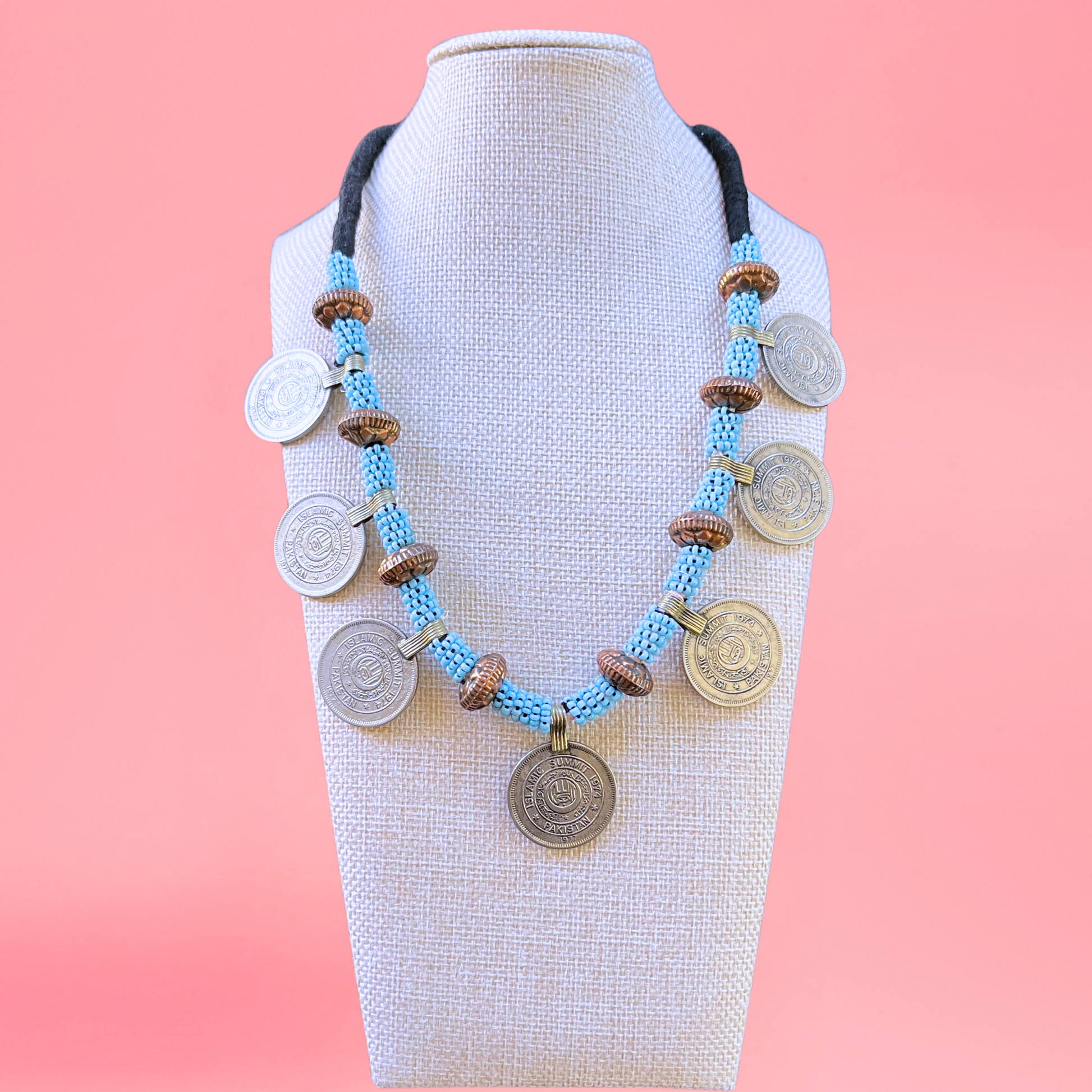 necklace modeled on a beige bust with a pink background. Necklace features persian coins, copper beads, and light blue beads. view from the front