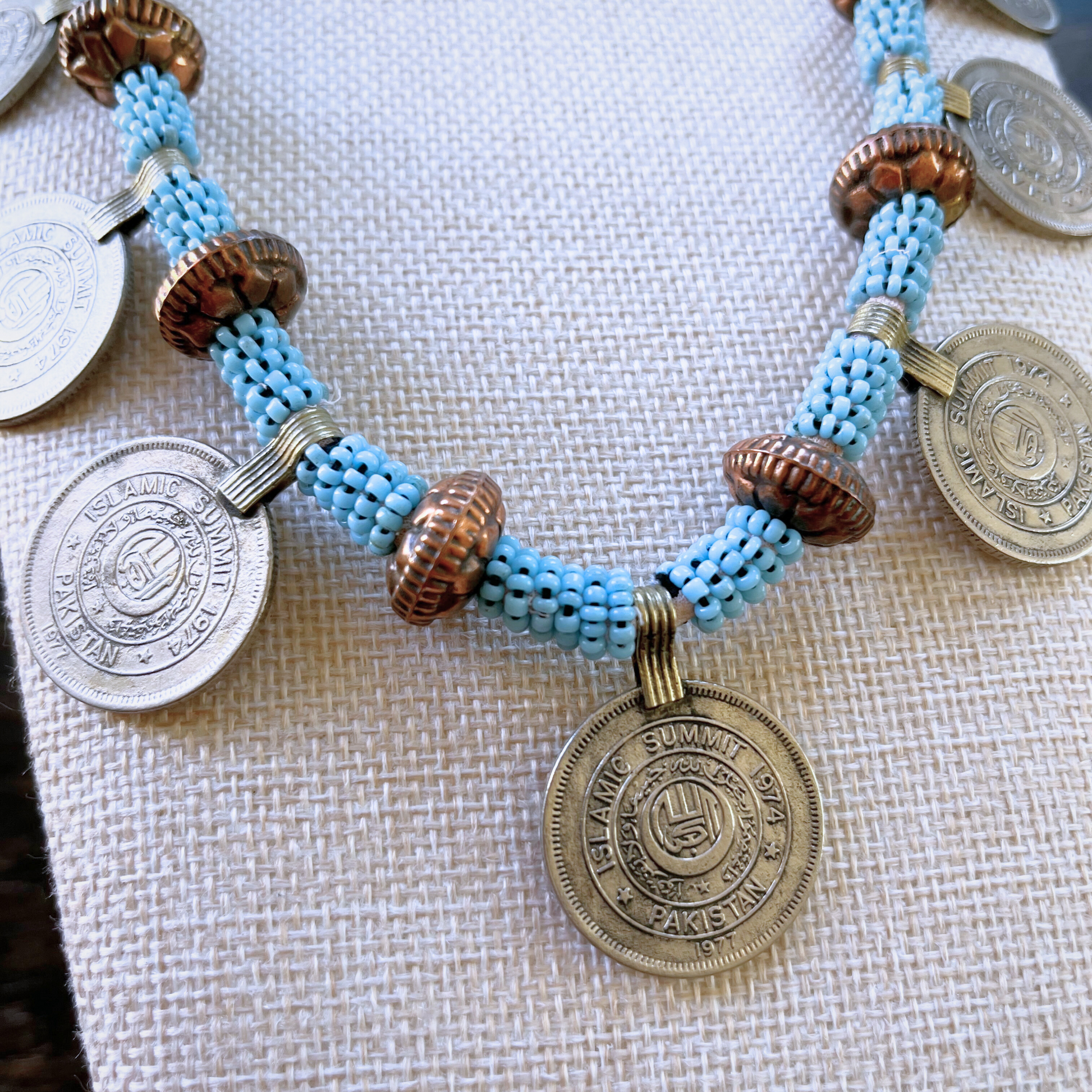 close-up view from above of a necklace modeled on a beige bust. Necklace features vintage persian coins, copper beads, and light blue beads.