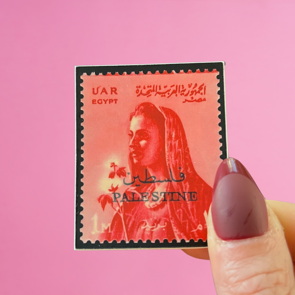 Front view of fingers holding a red Syria Egypt UAR stamp with an image of a woman and an overprint of a Palestine postmark. Against a pink background
