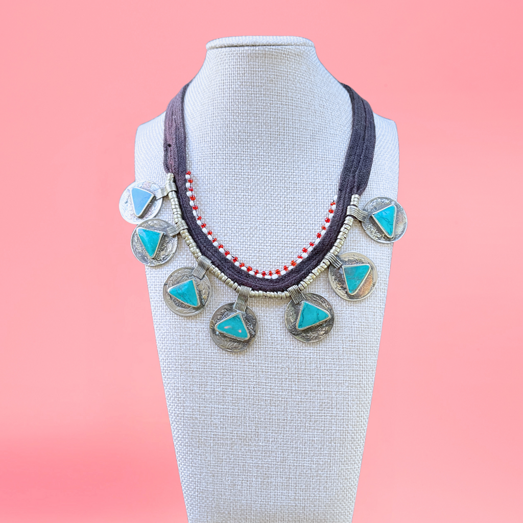 necklace modeled on a beige bust with a pink background. Necklace features persian coins, triangle-shaped turquoise stones, red and white beads. view from the front