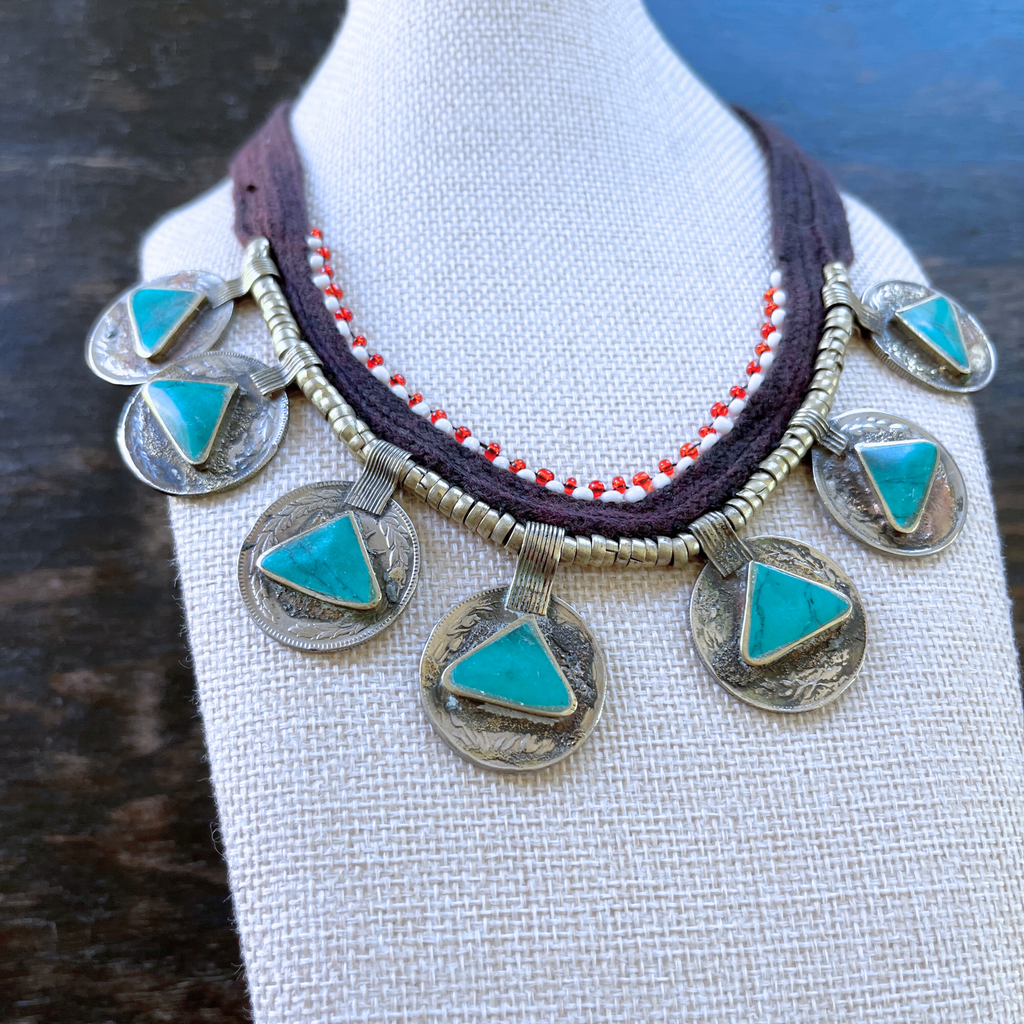 necklace modeled on a beige bust with a dark wood background. Necklace features persian coins, triangle-shaped turquoise stones, red and white beads. view from above