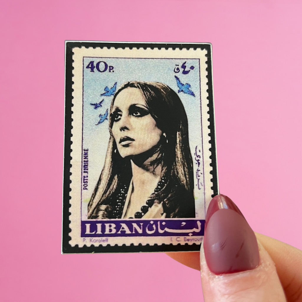 close-up view of fingers holding a sticker featuring a stamp issued by Lebanon and an image of Fairouz, the legendary Lebanese singer, with a pink background