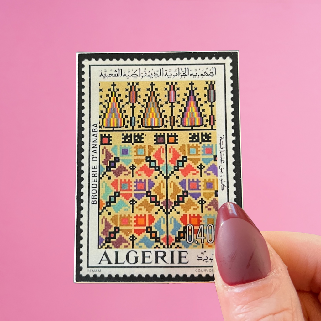Colorful Algerian postage stamp held by a hand against a pink background