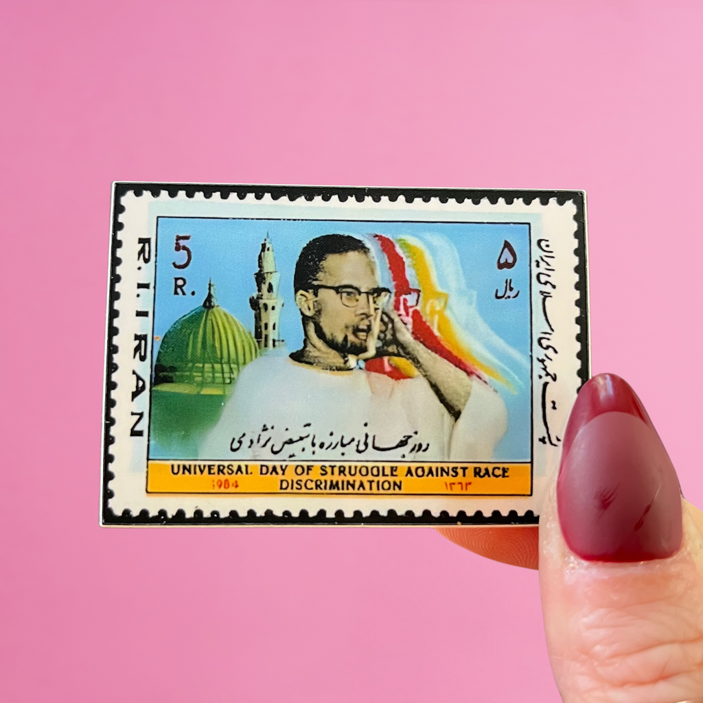 Front view of fingers holding an Iranian stamp with an image of Malcolm X in front of a mosque, against a pink background