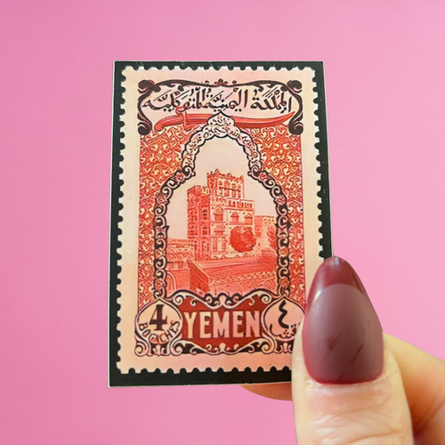 Front view of fingers holding a Yemen stamp with an image of Bab al-Yemen, the ancient gateway to Sana'a. Against a pink background