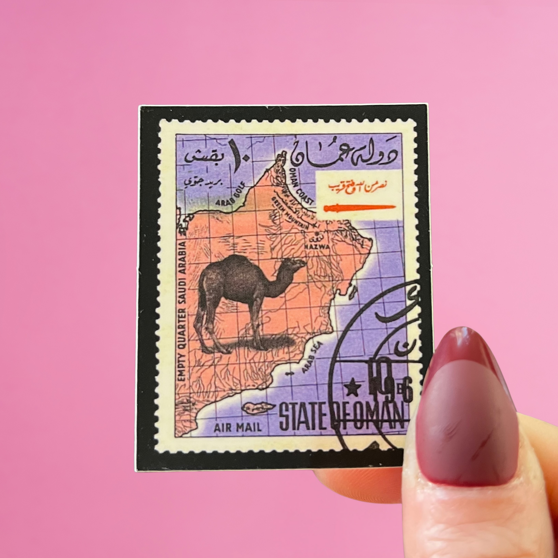 Front view of fingers holding an Oman stamp with an image of a camel on the map of Oman. Against a pink background