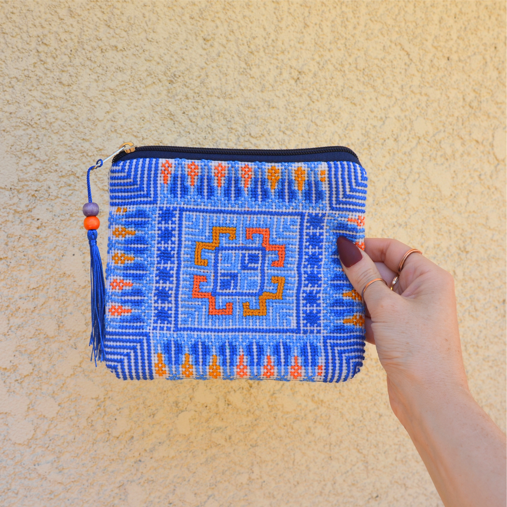 Palestinian tatreez embroidered purse/wallet/clutch bag. blue and orange cypress tree and moon with feathers design. held by a hand against a off-white wall. view from the front