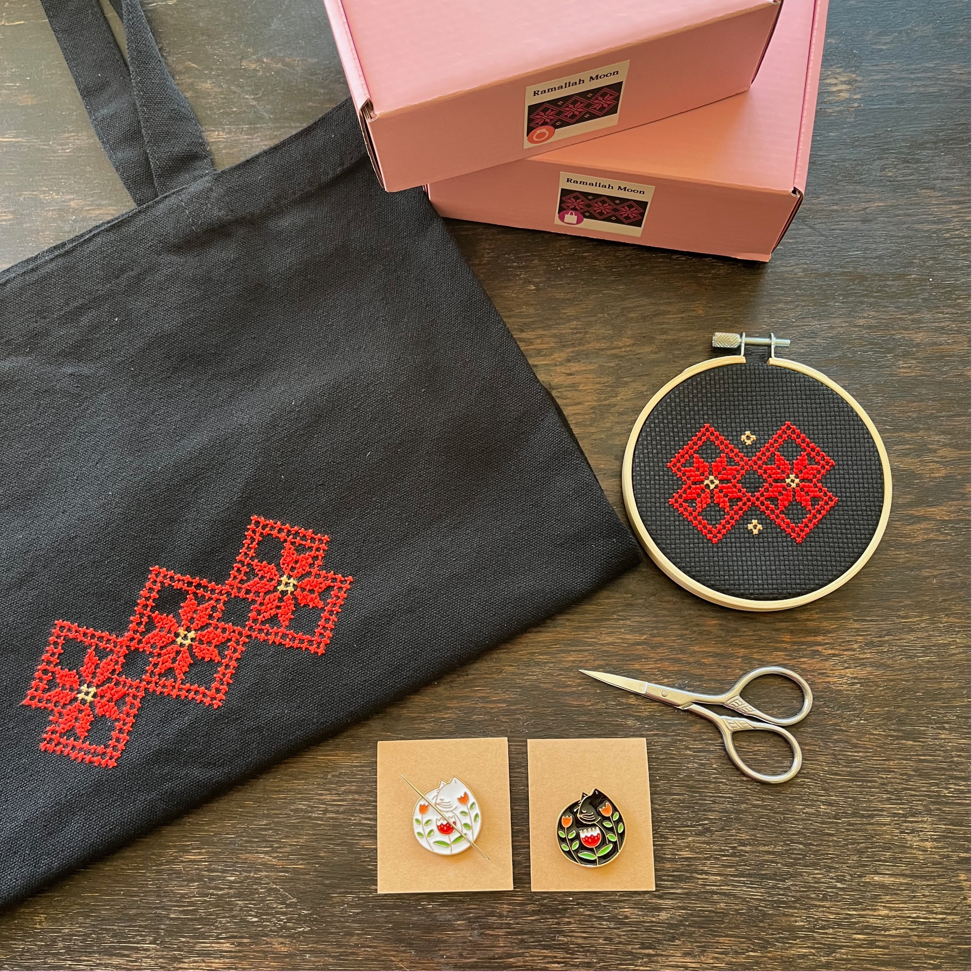 Tatreez tote bag kit and tatreez hoop kit with Ramallah Star Moon design. black tote bag, pink Rahaal tatreez kit box, cat needle minders, and embroidery scissors,  view from above