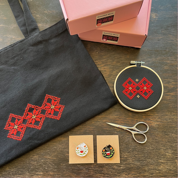 Tatreez tote bag kit and tatreez hoop kit with Ramallah Star Moon design. black tote bag, pink Rahaal tatreez kit box, cat needle minders, and embroidery scissors,  view from above