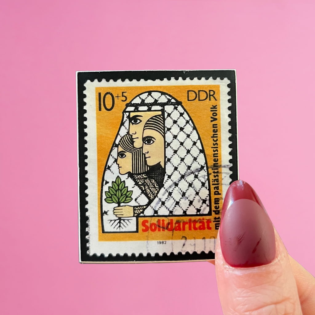 close-up of fingers holding an East Germany stamp stamp with an image of a Palestinian family wrapped in a kuffiyeh and holding an olive tree. against a pink background