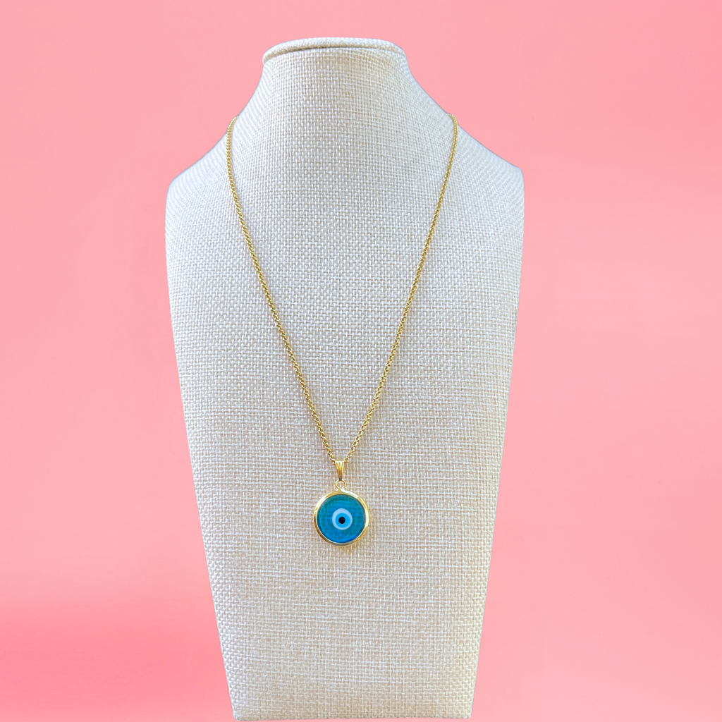 light blue handmade turkish evil eye necklace modeled on beige bust with pink background. lampwork glass evil eye framed in gold-plated stainless steal with gold-plated stainless steel chain. view from the front