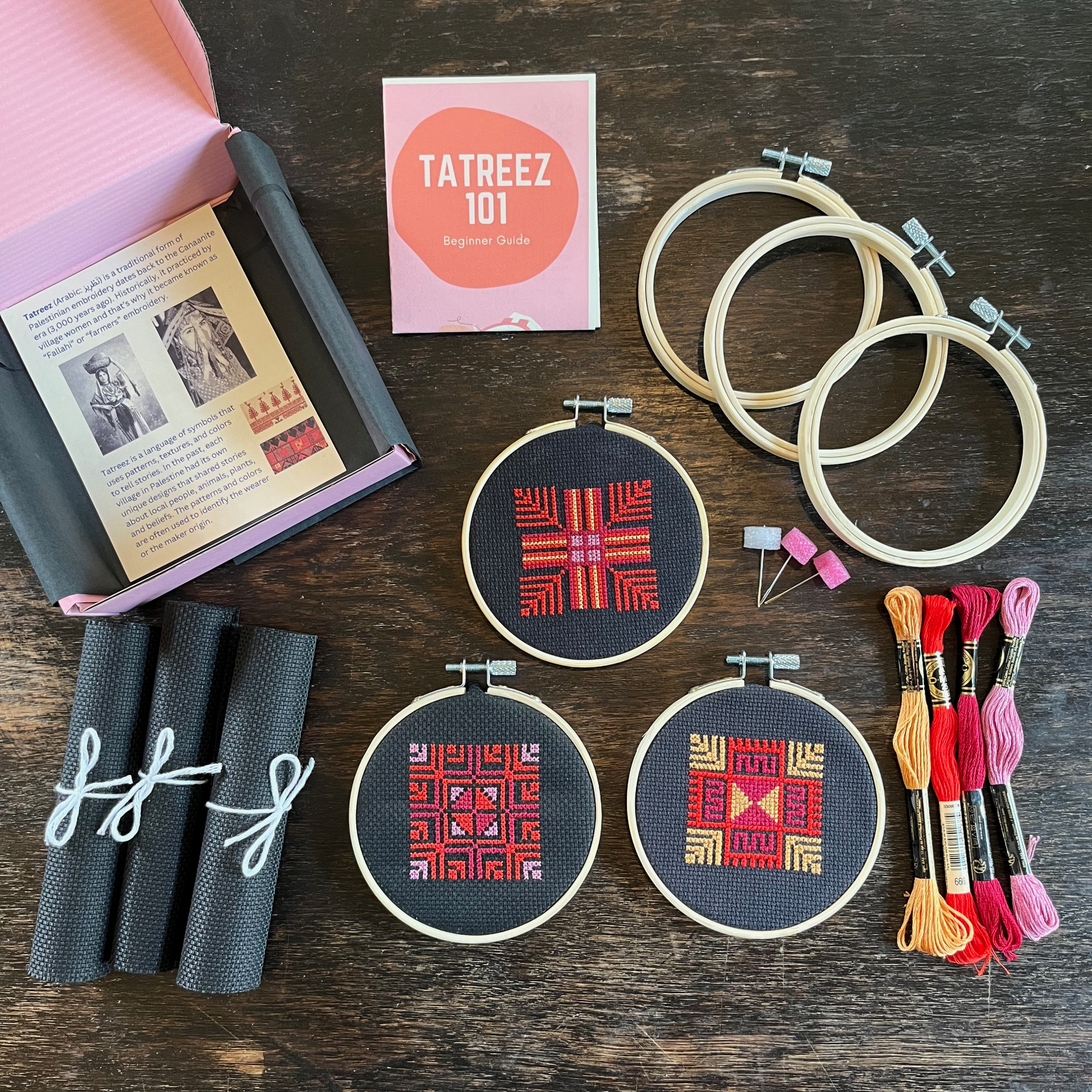 Tatreez kit with materials laid out, including black aida cloths, tatreez instructions, moon with feathers tatreez patterns, embroidery threads, embroidery needles, and embroidery hoops. view from above