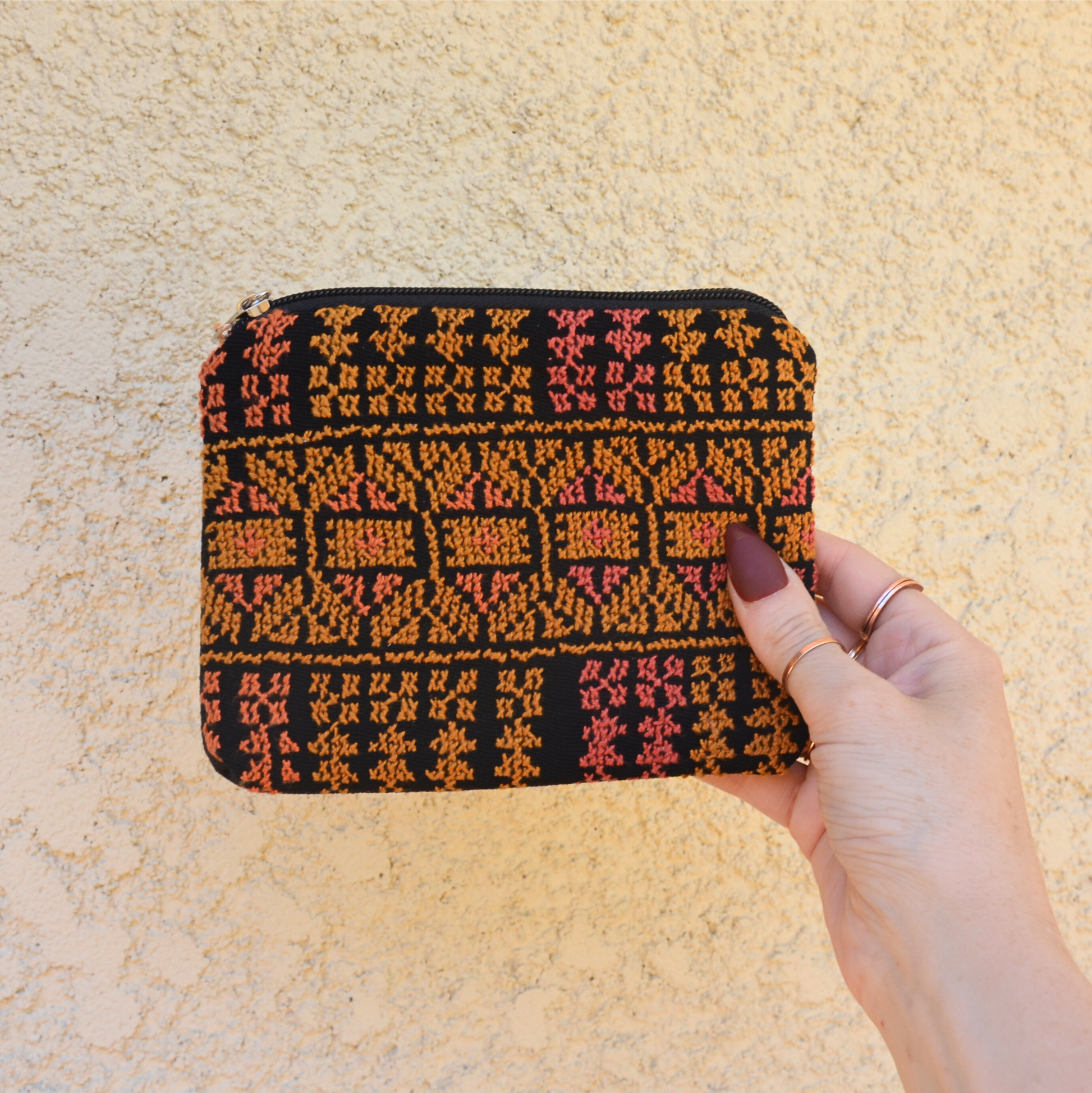 Zippered pouch/wallet with pink and gold handmade Palestinian tatreez embroidery. held by a hand against an off-white background, view from the front