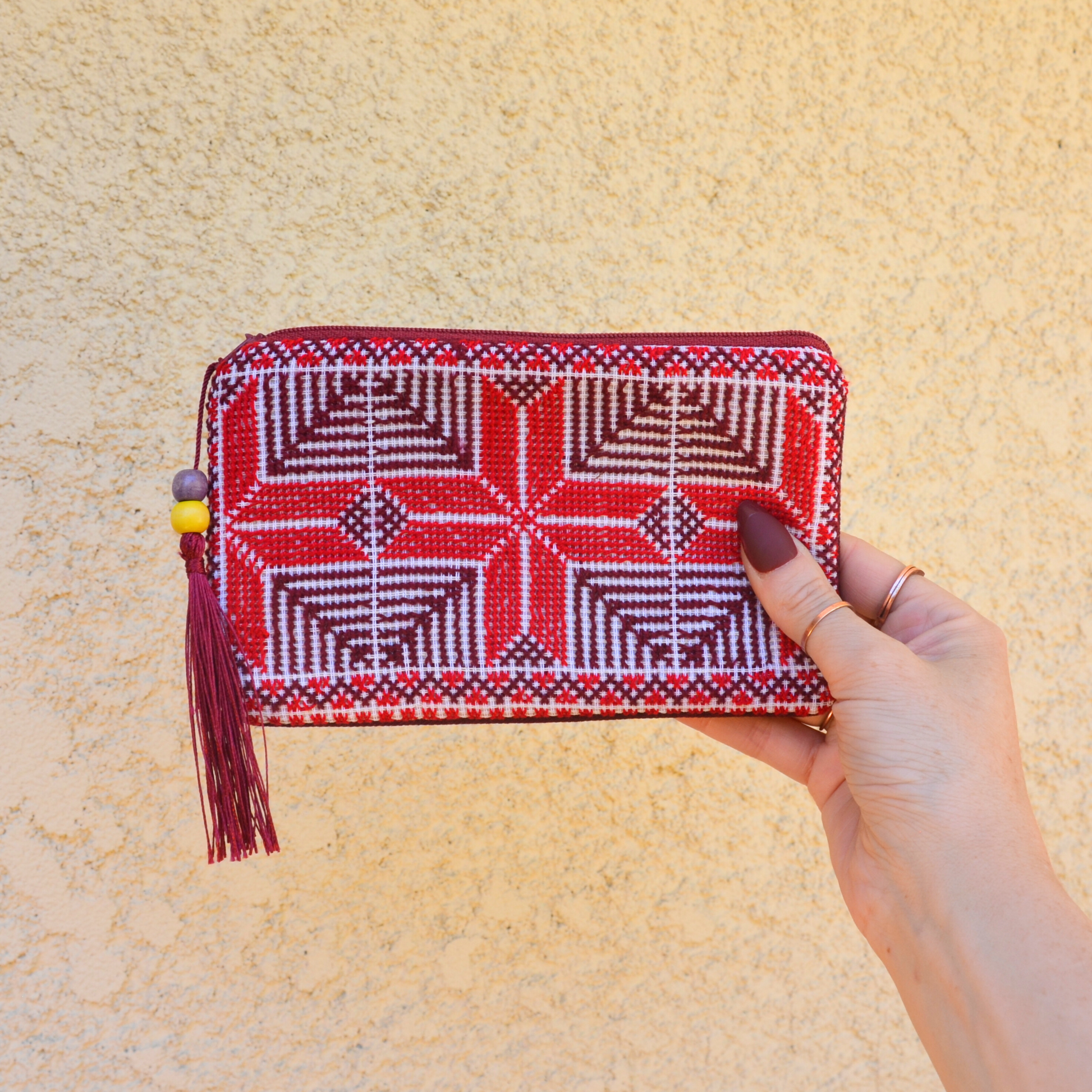 Palestinian tatreez embroidered purse/wallet/clutch bag. red and purple moon with feathers design. held by a hand against a off-white wall. view from the front