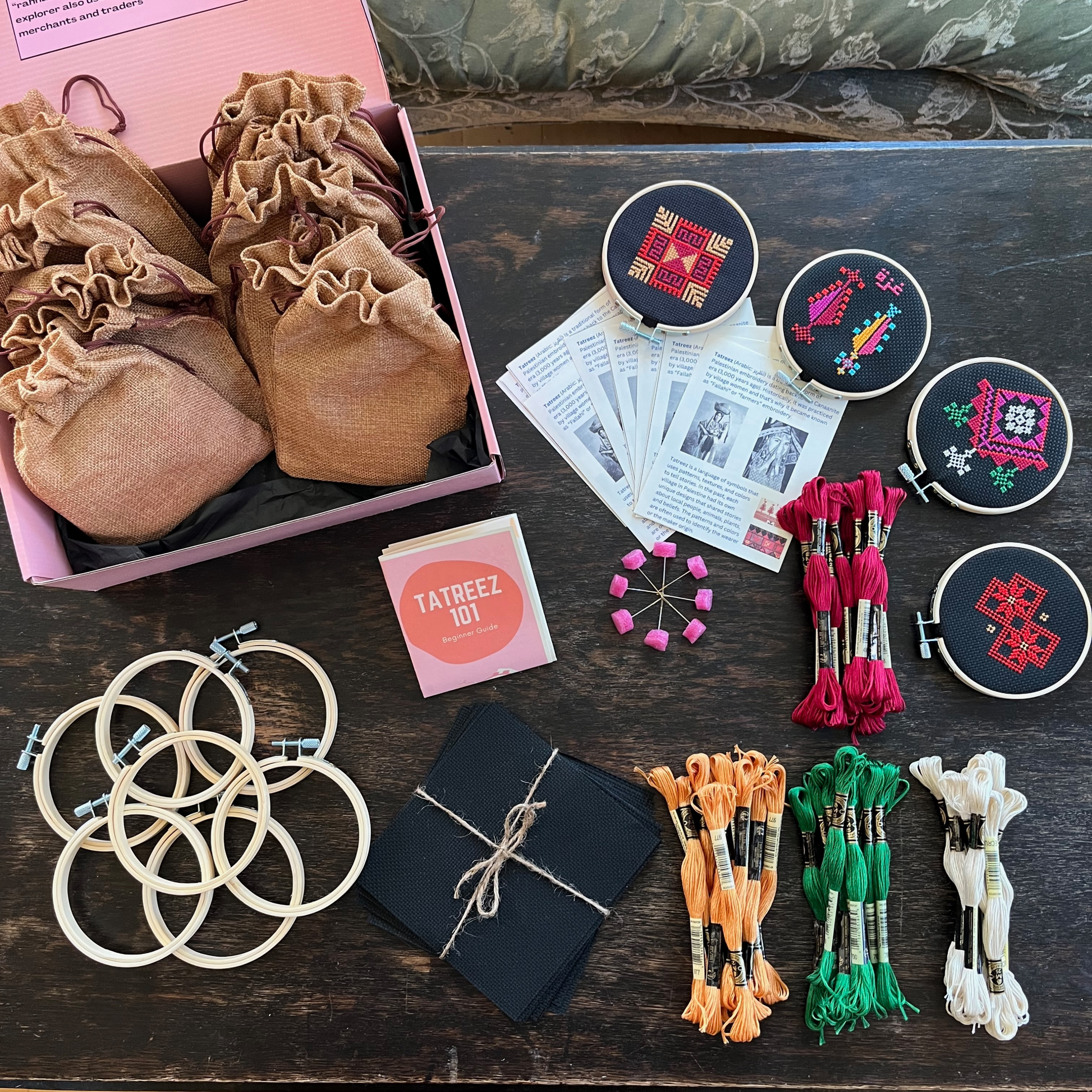 Tatreez workshop kit with materials laid out, including black aida cloth, tatreez instructions, tatreez patterns, embroidery threads, embroidery needle and pins, and embroidery hoops. view from above