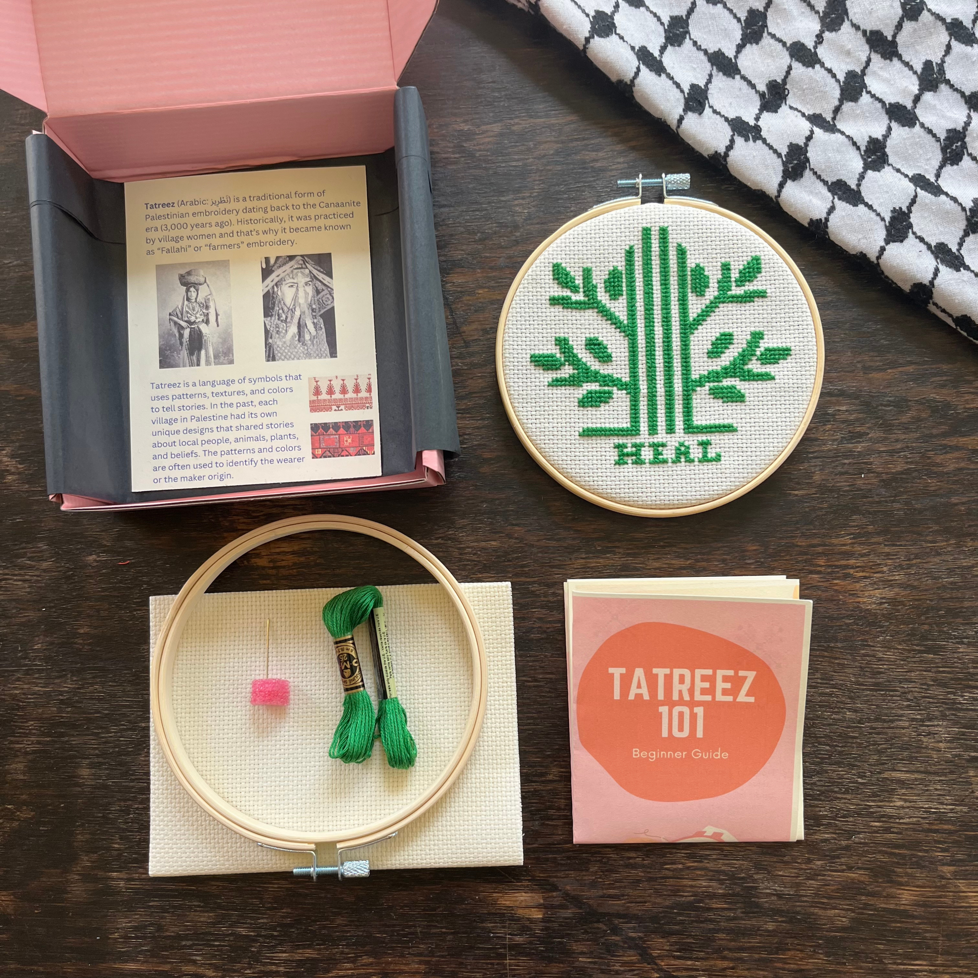 Tatreez kit with materials laid out, including white aida cloths, tatreez instructions, Heal Palestine olive tree tatreez patterns, embroidery thread, embroidery needle, and embroidery hoop. view from above