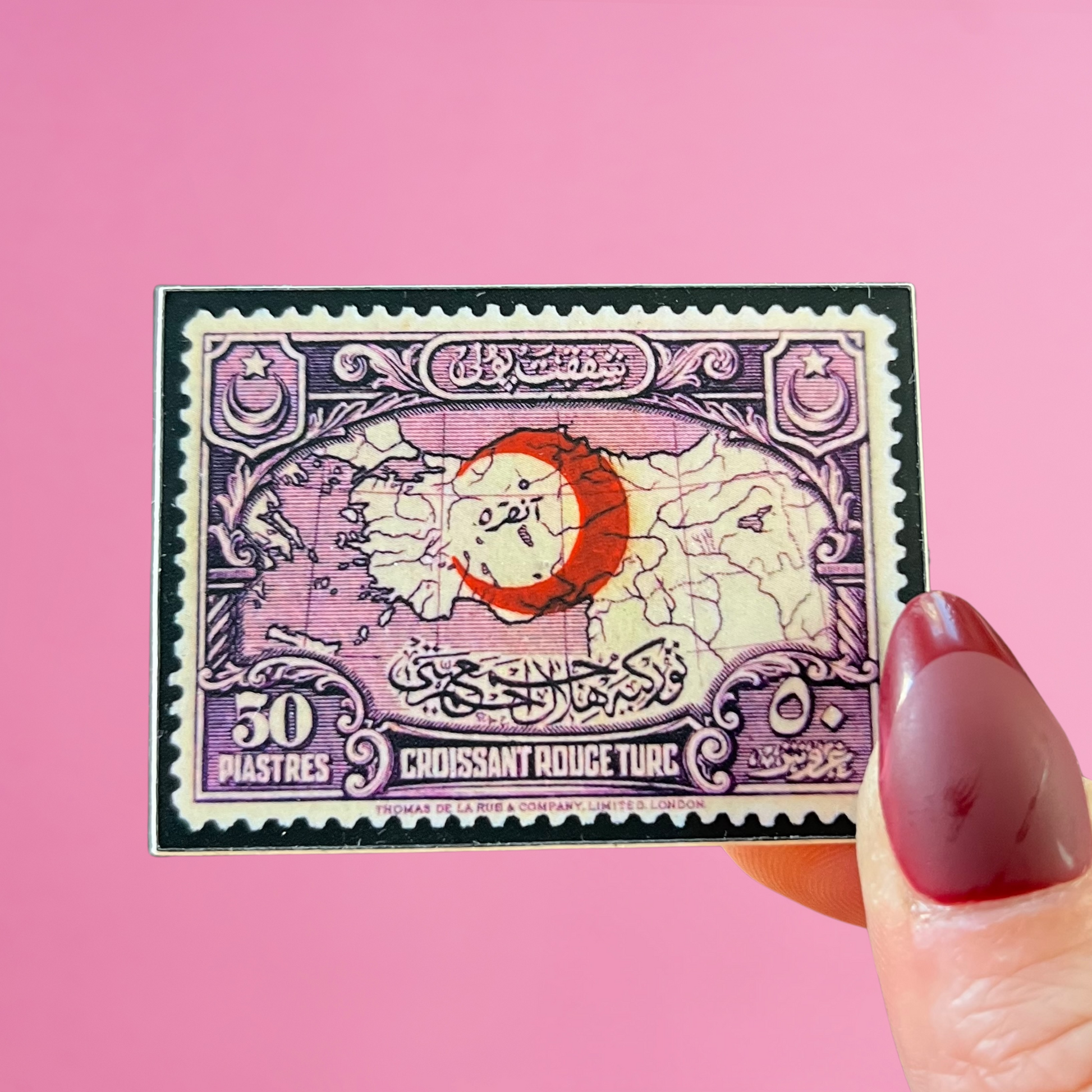 close up of fingers holding a Turkish stamp sticker featuring a purple map of the country with a red crescent moon. against a pink background.