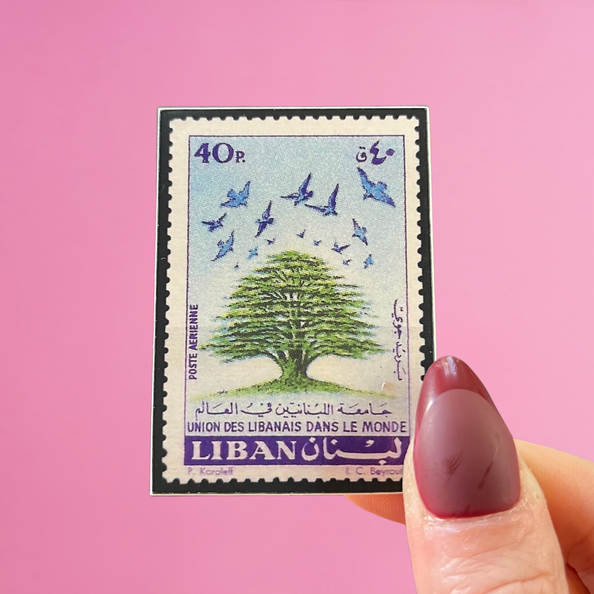 Front view of fingers holding a blue and green Lebanon stamp with an image of a Cedar tree. Against a pink background