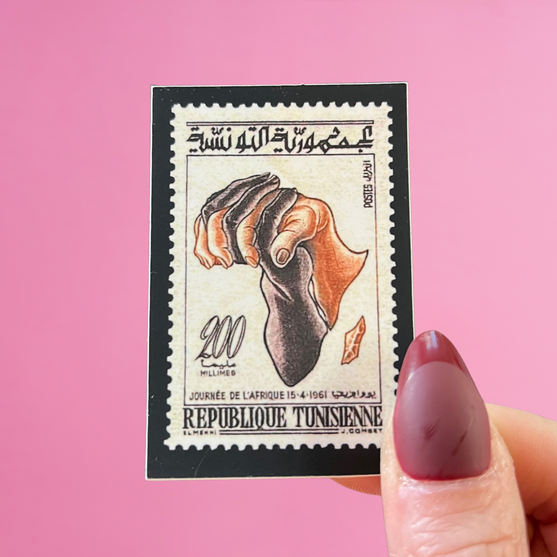 Front view of fingers holding a Tunisia stamp with an image of the map of Africa with Black and Brown hands embracing. Against a pink background