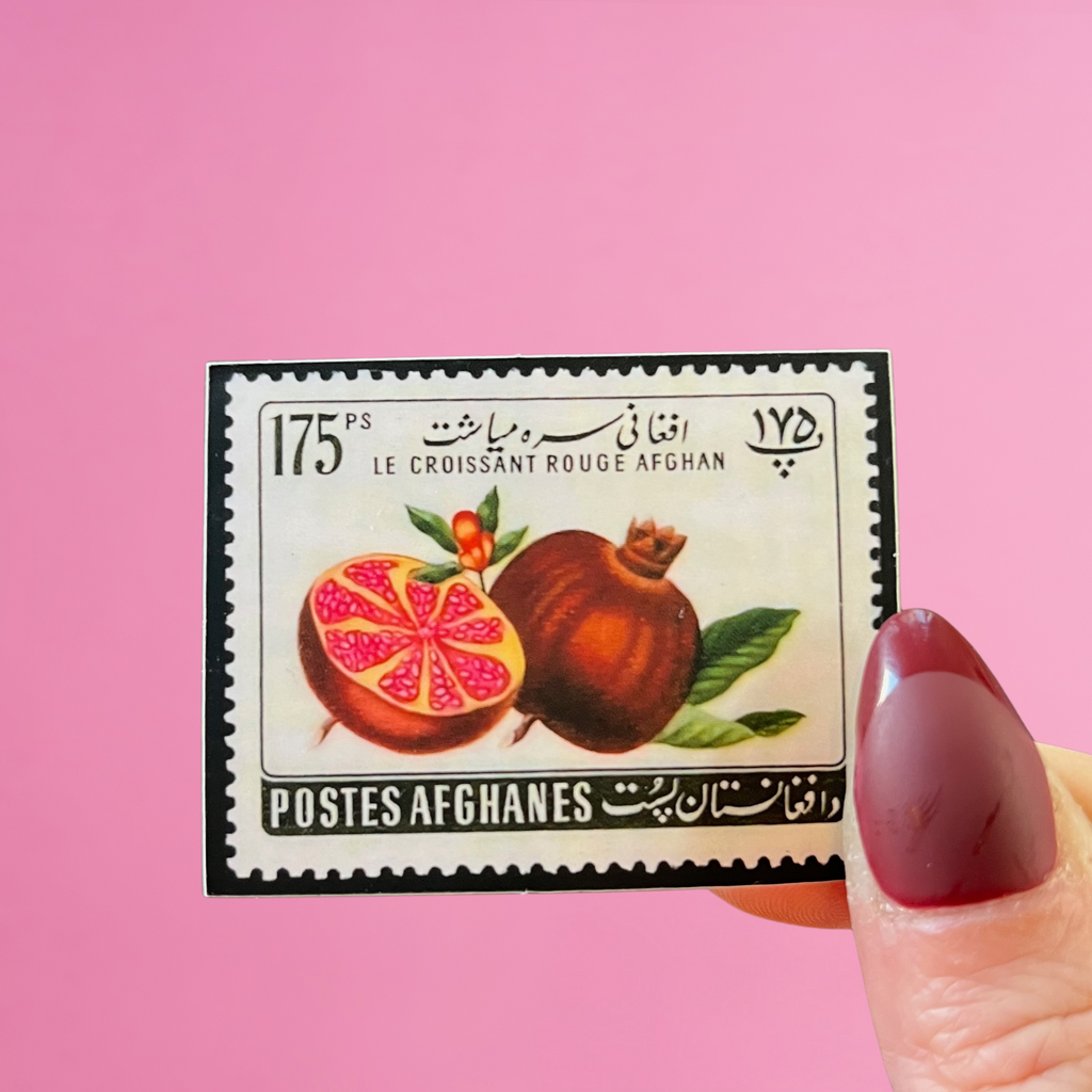 close-up front view of fingers holding an Afghanistan stamp sticker with an image of two pomegranates. pink background