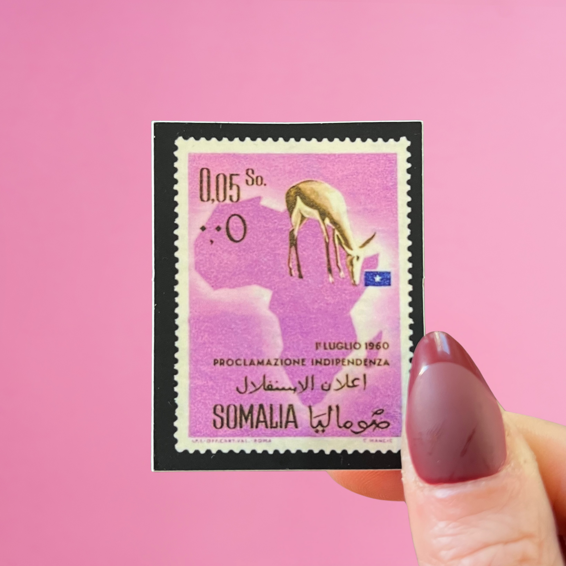 Front view of fingers holding a purple Somalia stamp with an image of a deer on the map of Africa. Against a pink background
