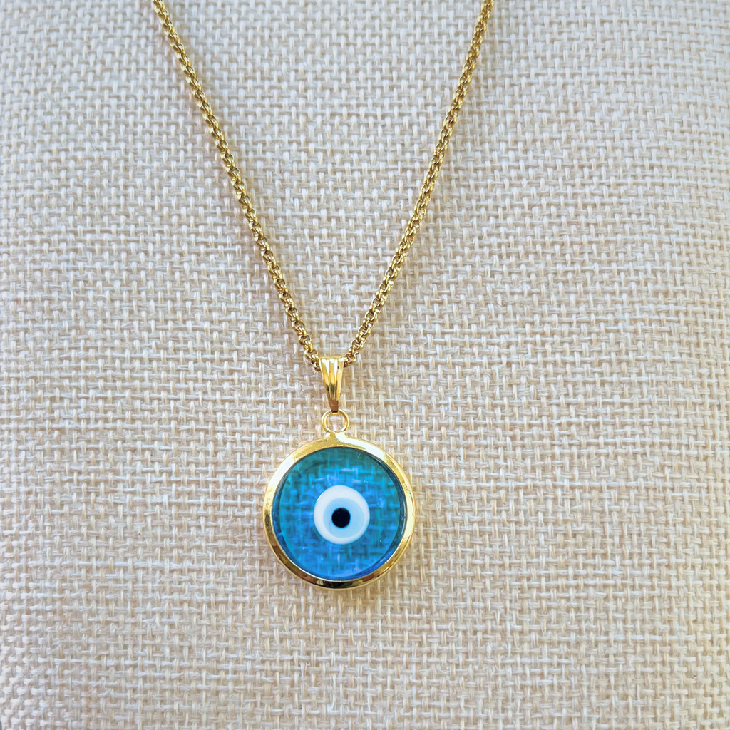 close-up frontal view of light blue handmade turkish evil eye necklace modeled on beige bust. lampwork glass evil eye framed in gold-plated stainless steal with gold-plated stainless steel chain.
