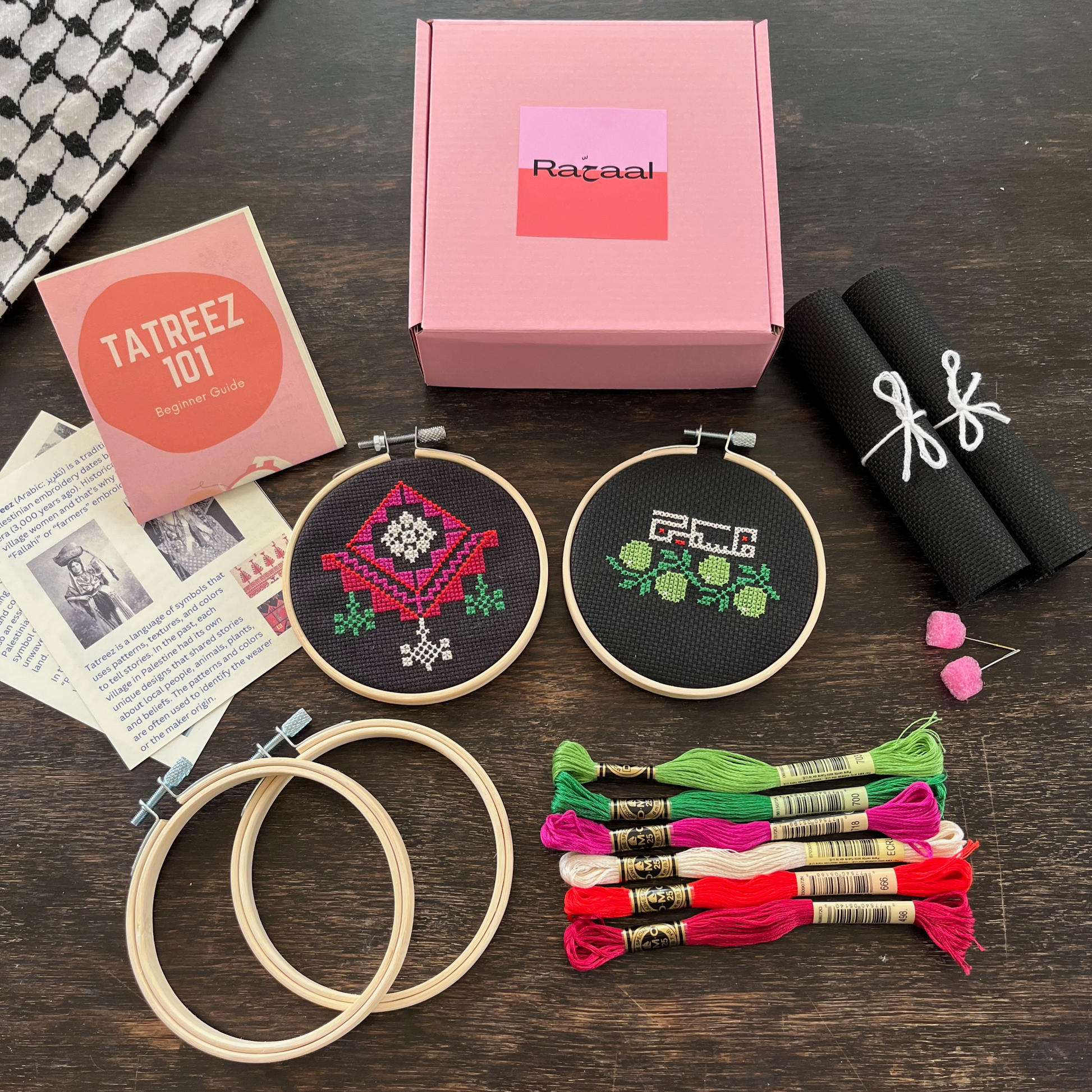 Tatreez kit with materials laid out, including black aida cloths, tatreez instructions, chandelier and olive & Palestine tatreez patterns, embroidery threads, embroidery needles, and embroidery hoops. view from above