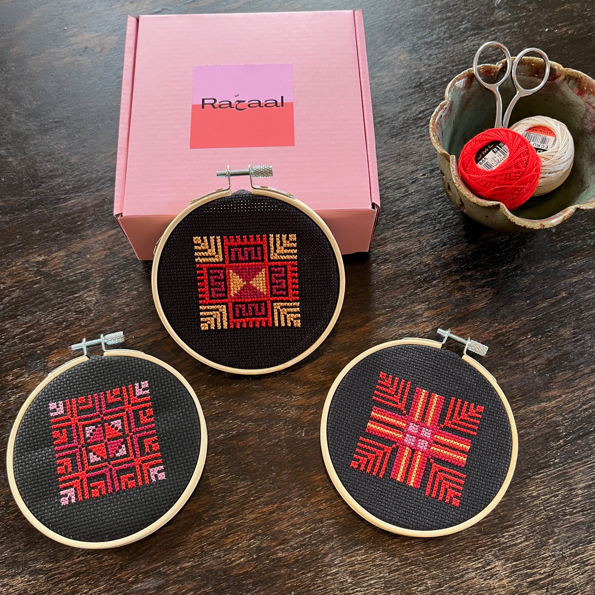Tatreez kit with three moon with feathers designs. black aida cloths, embroidery hoops, pink Rahaal tatreez kit box, embroidery scissors, and embroidery threads, view from above