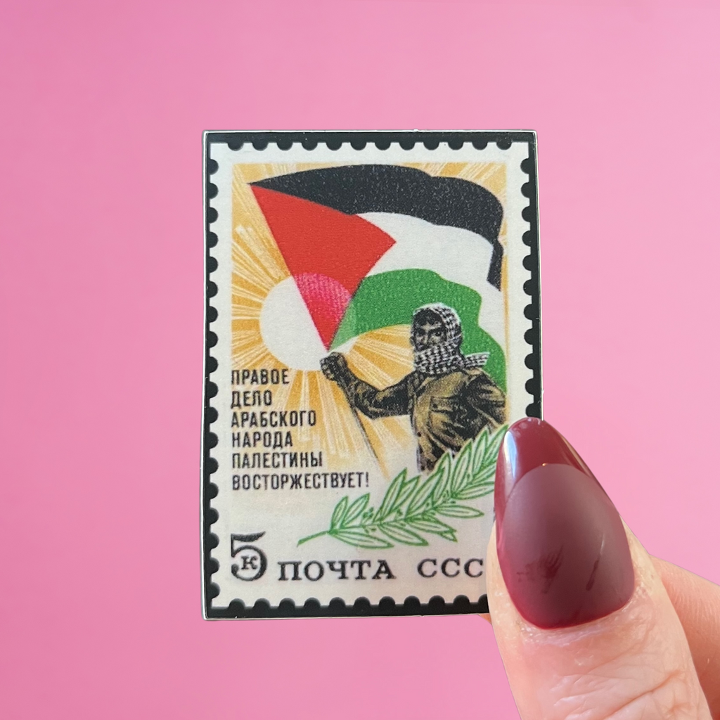 Front view of fingers holding a USSR stamp with an image of a Palestinian man wearing a kuffiyeh, an olive branch, and the Palestinian flag. Against a pink background