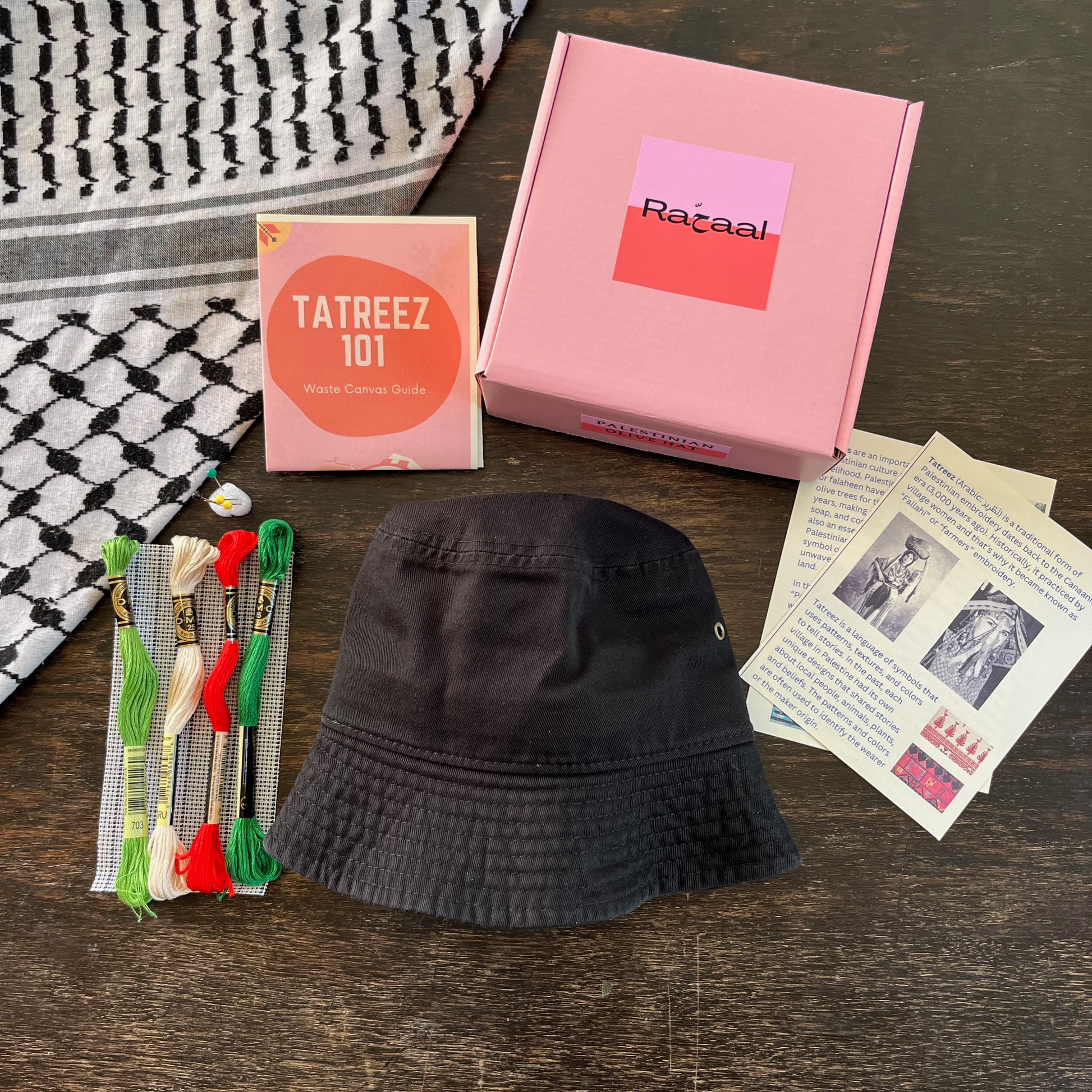 Tatreez bucket hat kit with materials laid out, including black bucket hat, tatreez instructions, olive and Palestine tatreez pattern, embroidery threads, embroidery needle and pins, and waste canvas. view from above