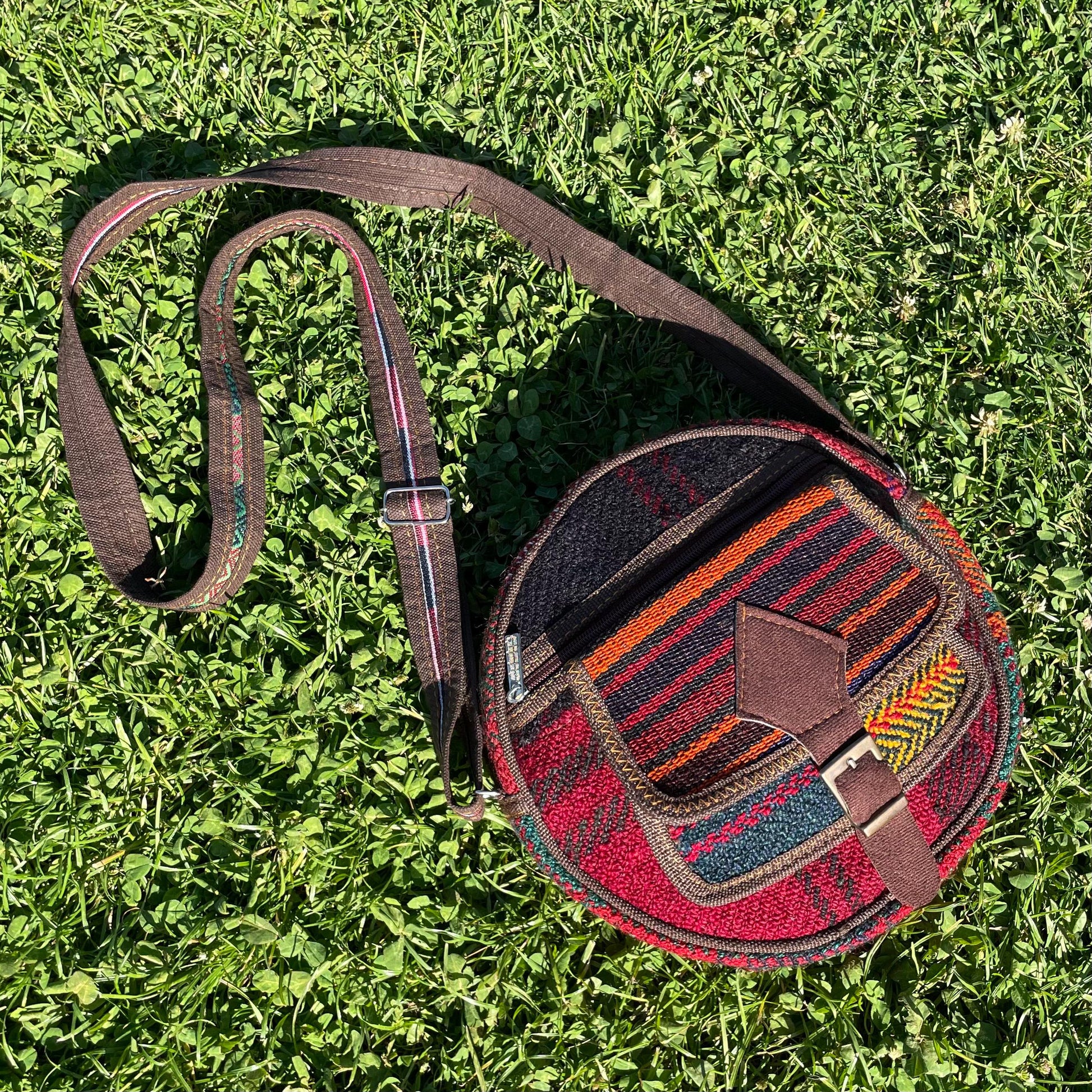 view from above of a colorful circle bag on a grass background. the purse has handwoven sections of wool, trimmed with brown fabric. The bag has a long strap, extra pockets, zippers, and a faux buckle over a snap closure.