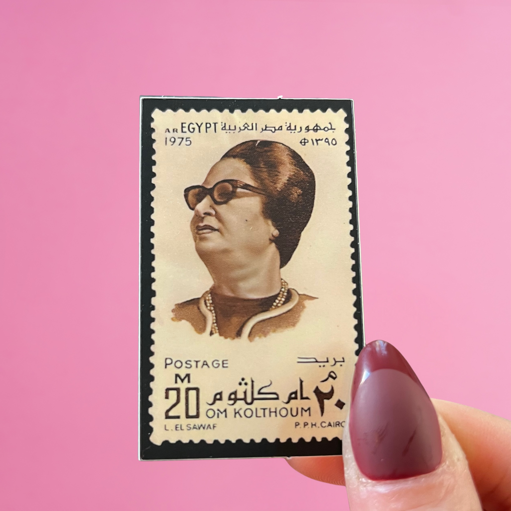 close up view of fingers holding an egyptian stamp with an image of Um Kalthoum, the legendary Egyptian singer, on a pink background