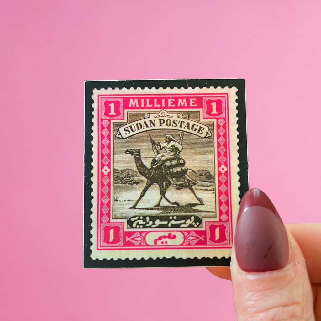 Front view of fingers holding a pink Sudan stamp with an image of a postman delivering mail on camelback. Against a pink background