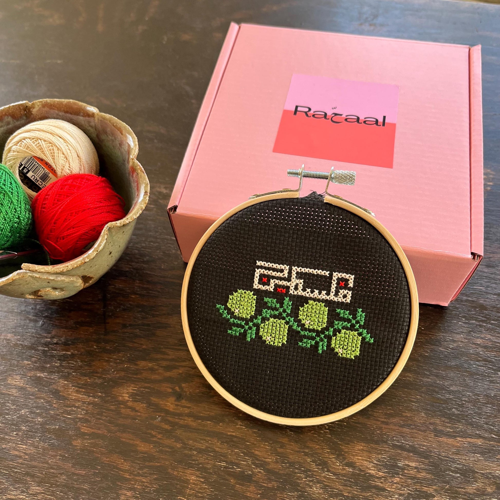 Tatreez kit with olive branch and Palestine design. black aida cloth, embroidery hoop, pink Rahaal tatreez kit box and embroidery threads, front view