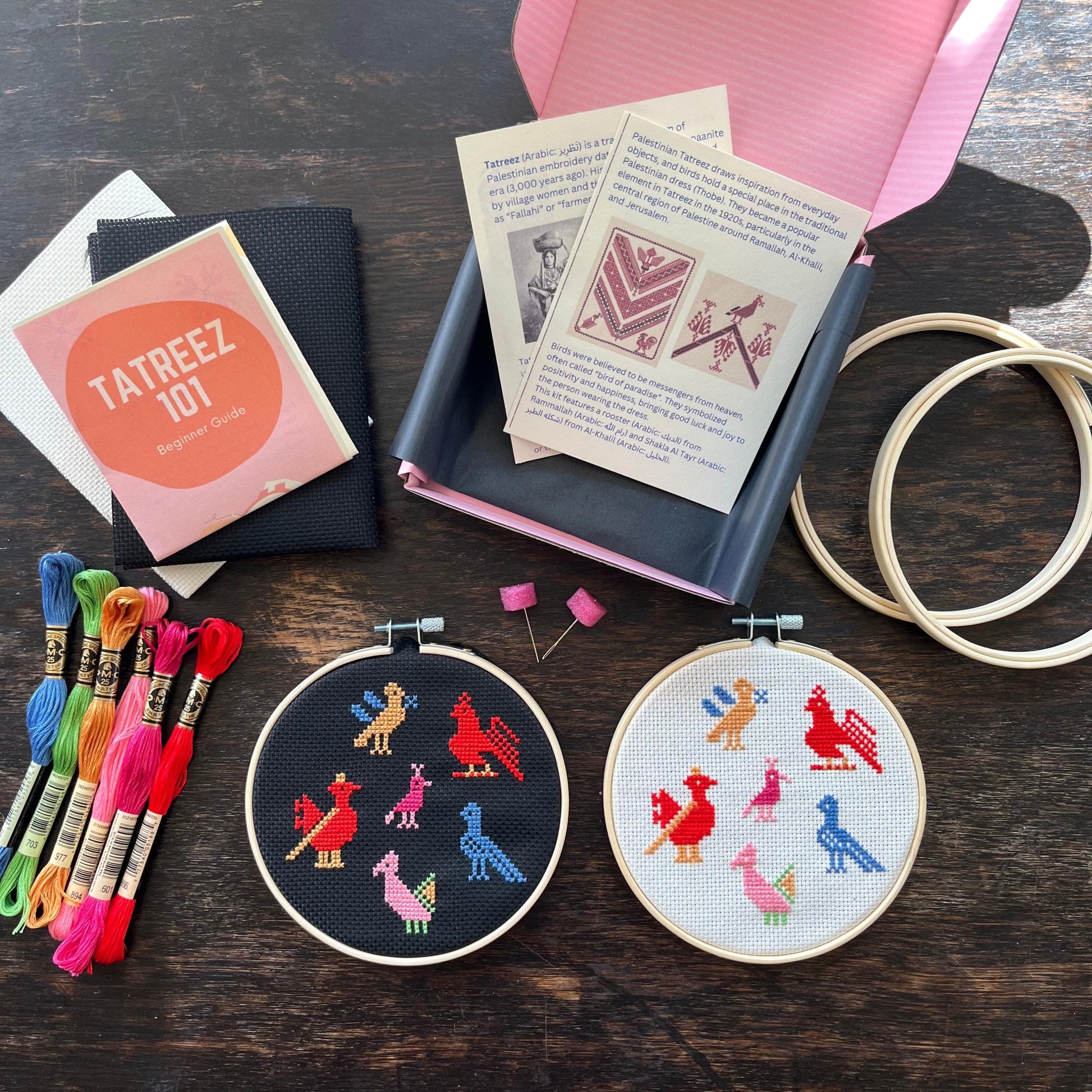 Tatreez kit with materials laid out, including black and white aida cloths, tatreez instructions, birds of Ramallah tatreez patterns, embroidery threads, embroidery needles, and embroidery hoops. view from above