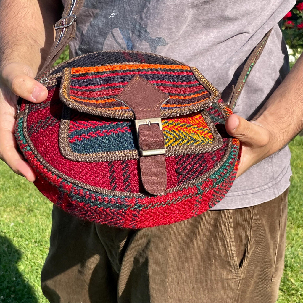 close-up view of a colorful circle bag handwoven sections of wool, trimmed with brown fabric. The bag has a long strap, front pockets, zippers, and a faux buckle over a snap closure