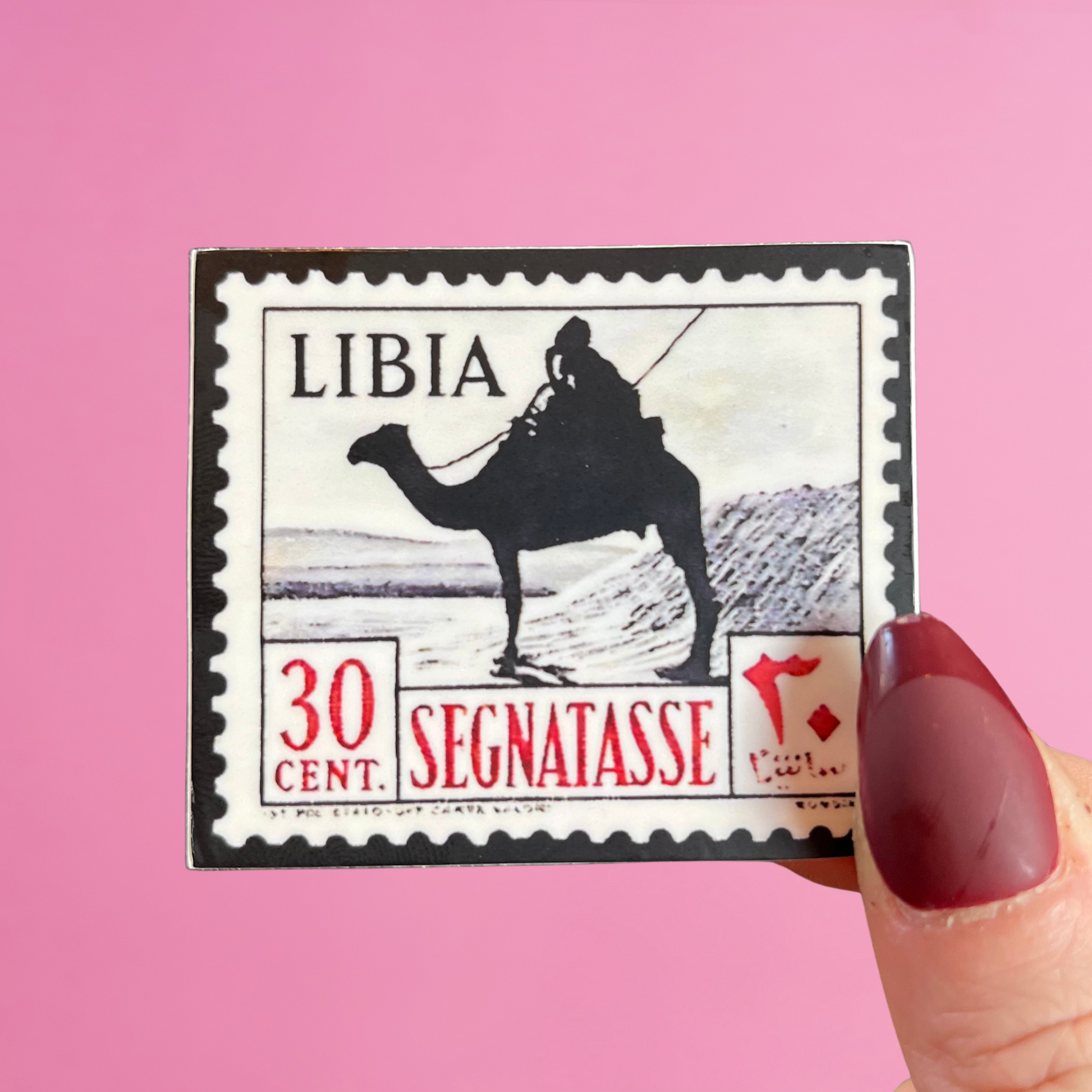 Front view of fingers holding a Libya stamp with an image of a camel and a rider in the desert. Against a pink background
