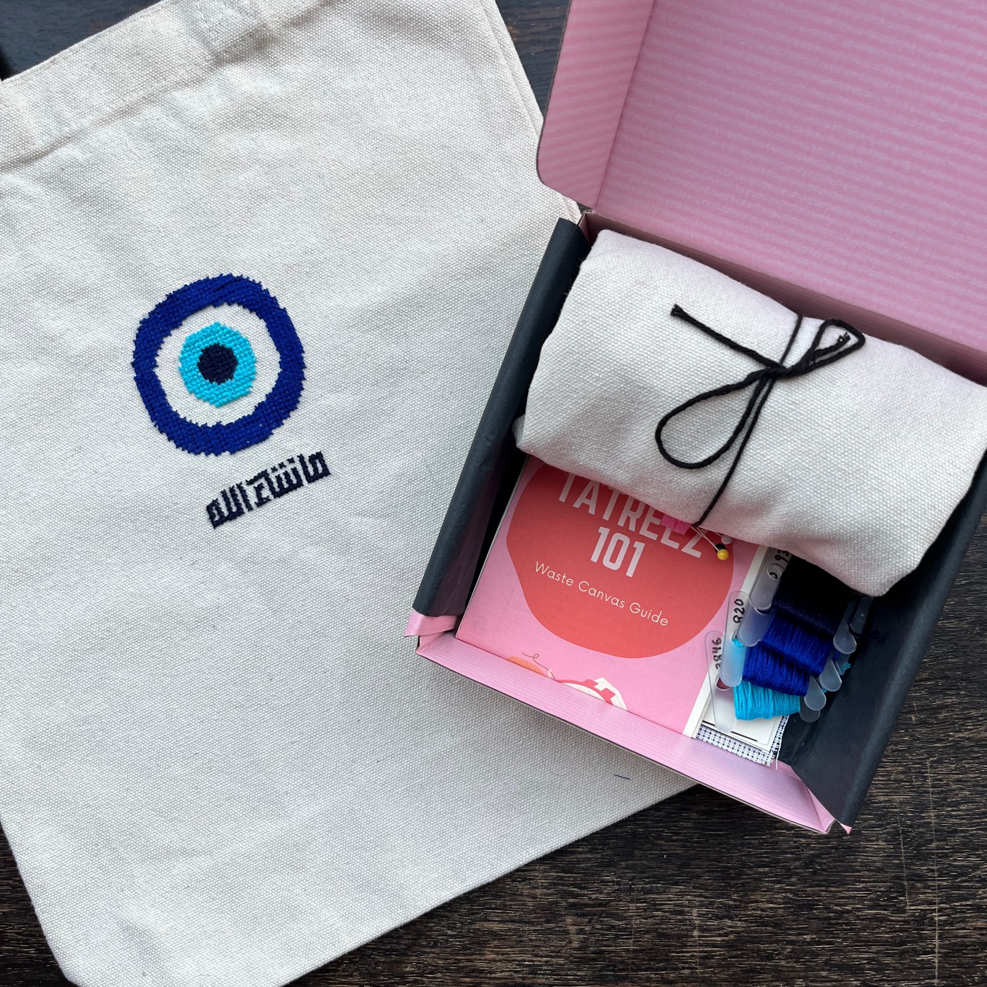 Tatreez tote bag kit with evil eye design. beige tote bag, pink Rahaal tatreez kit box with embroidery threads and tatreez instructions for beginners. view from above