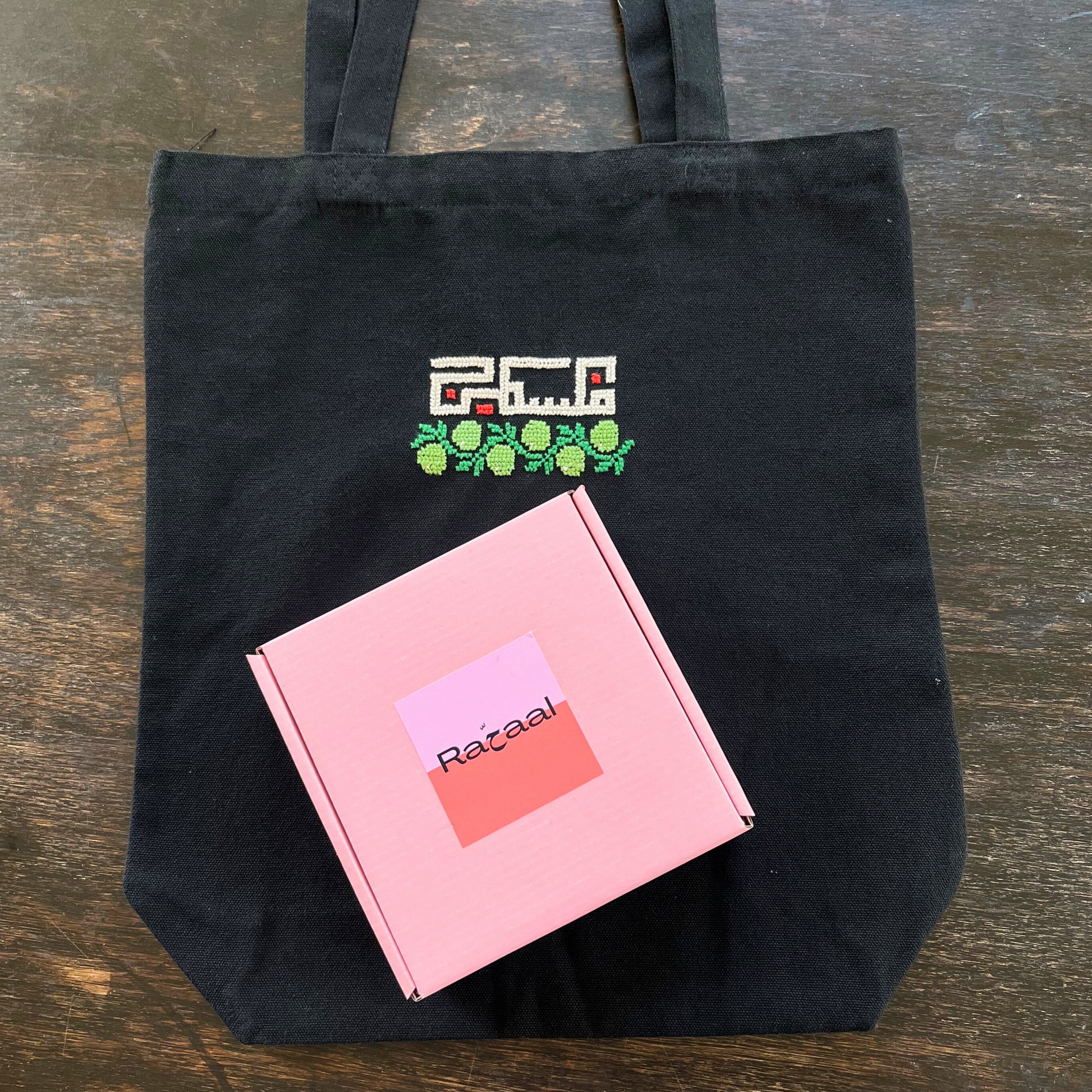 Tatreez tote bag kit with olive design. black tote bag and pink Rahaal tatreez kit box, view from above