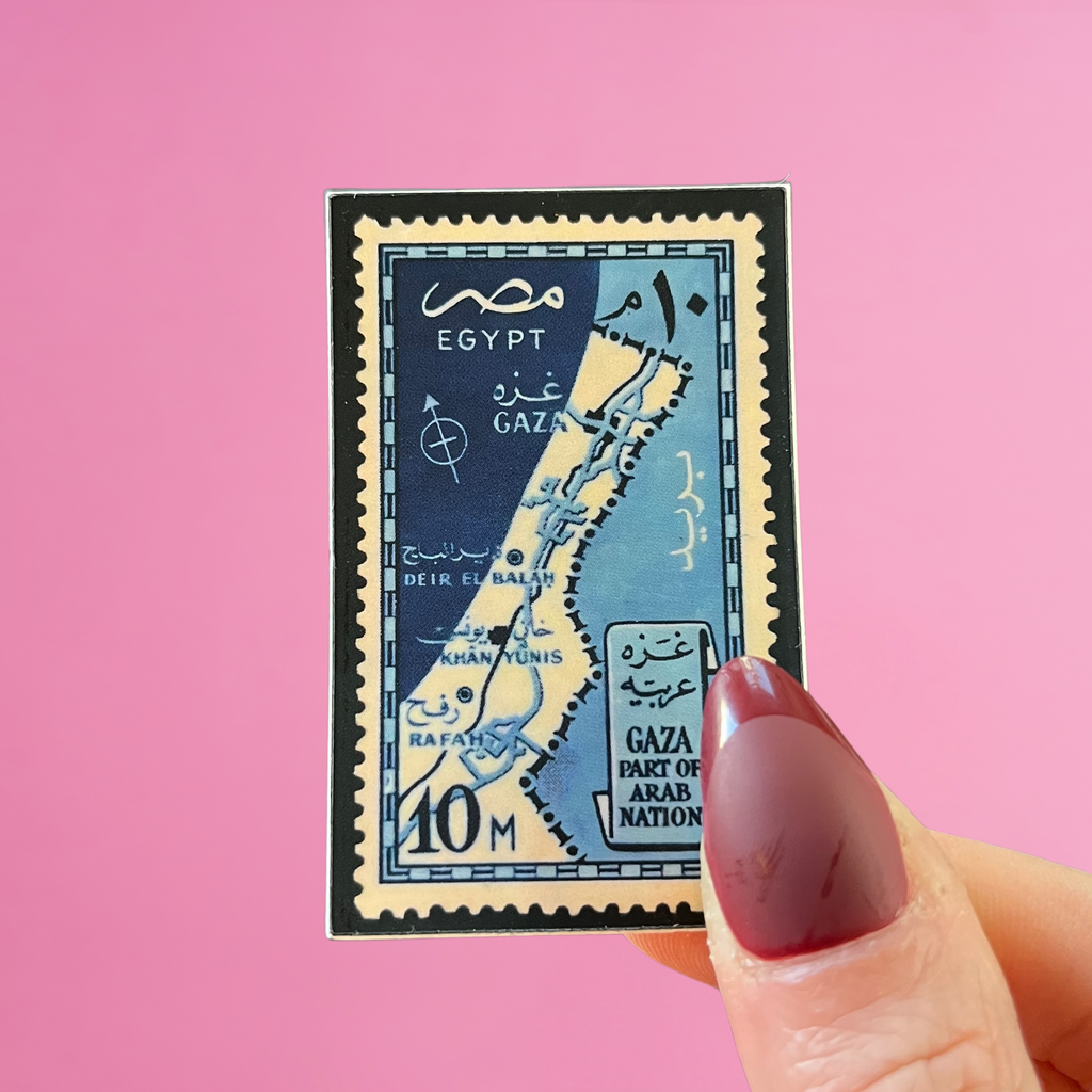 Front view of fingers holding a blue Egypt stamp with an image of Gaza in Palestine. Against a pink background