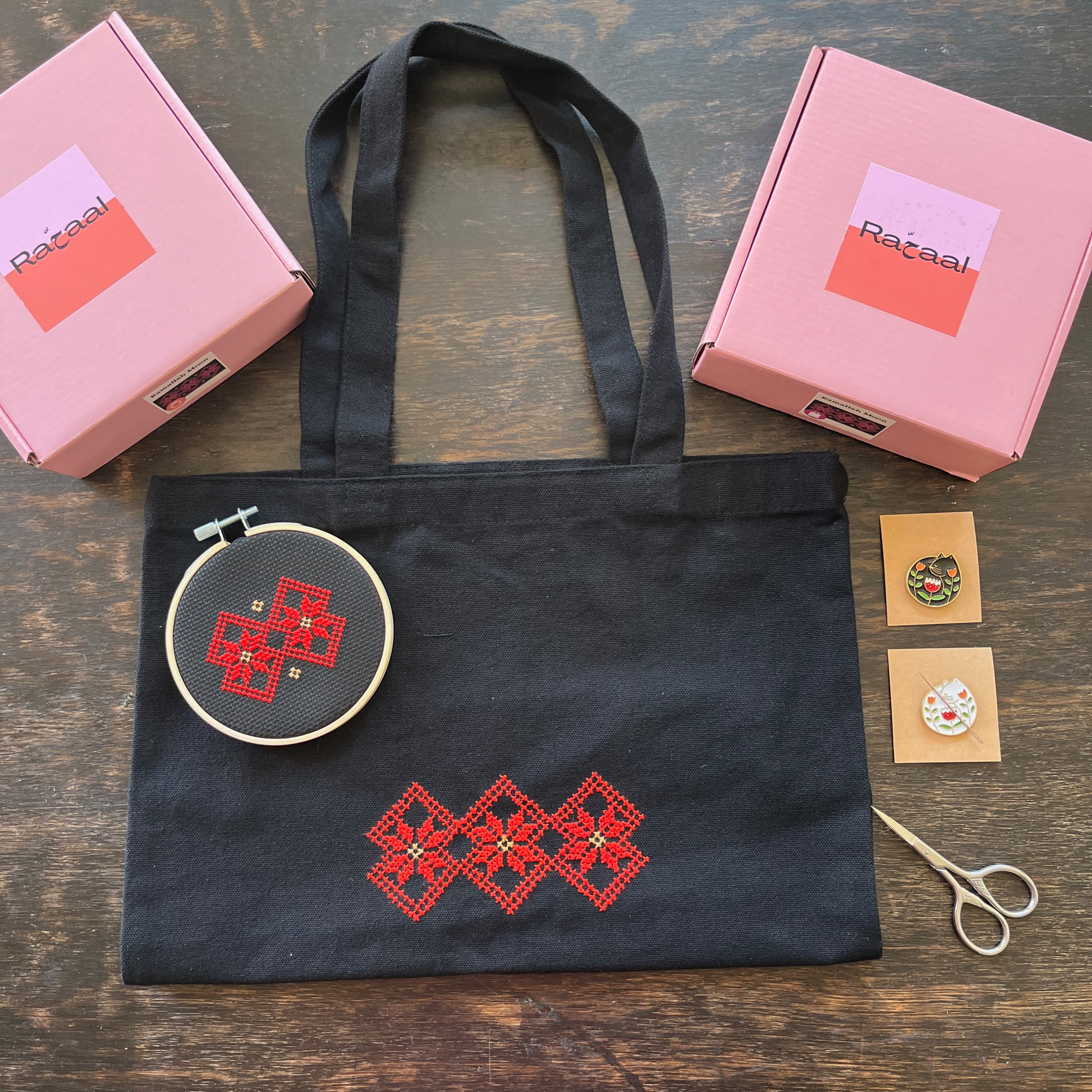 Tatreez tote bag kit and tatreez hoop kit with Ramallah Star Moon design. black tote bag, pink Rahaal tatreez kit box, cat needle minders, and embroidery scissors,  view from above