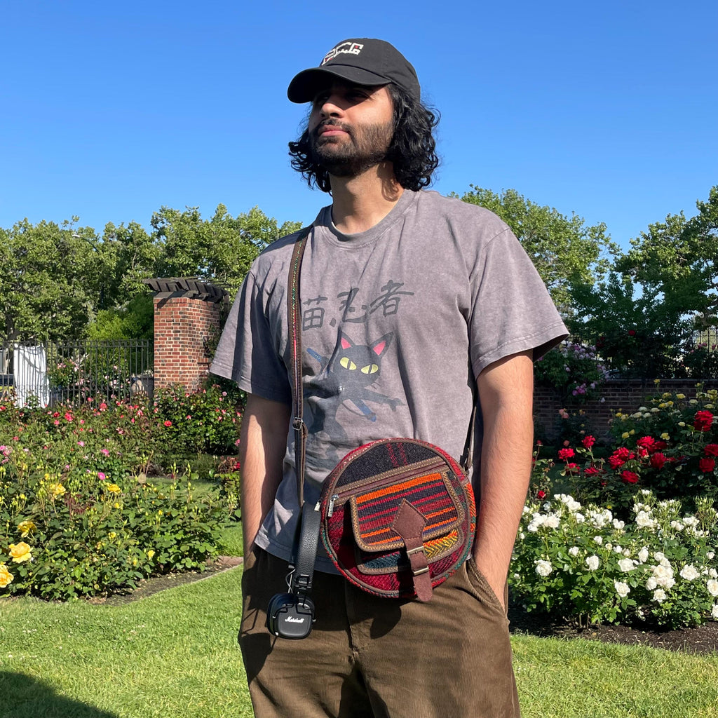 view from the front of a colorful circle bag modeled on a man in a garden. the purse has handwoven sections of wool, trimmed with brown fabric. The bag has a long strap, front pockets, zippers, and a faux buckle over a snap closure