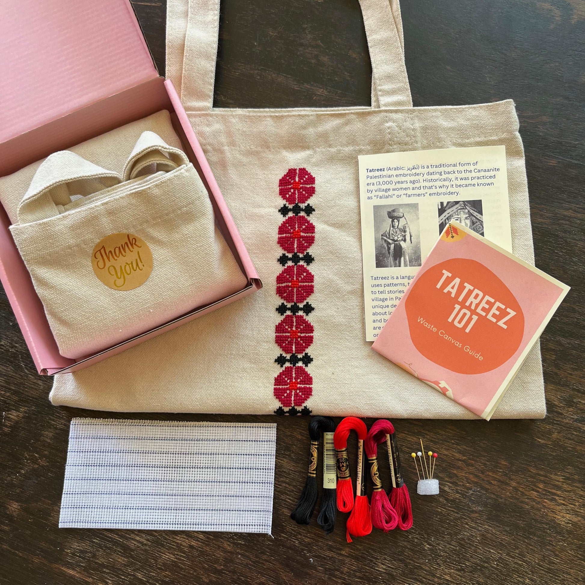 Tatreez tote bag kit with materials laid out, including beige tote bag, tatreez instructions, Ramallah flower tatreez pattern, embroidery threads, embroidery needle and pins, and waste canvas. view from above