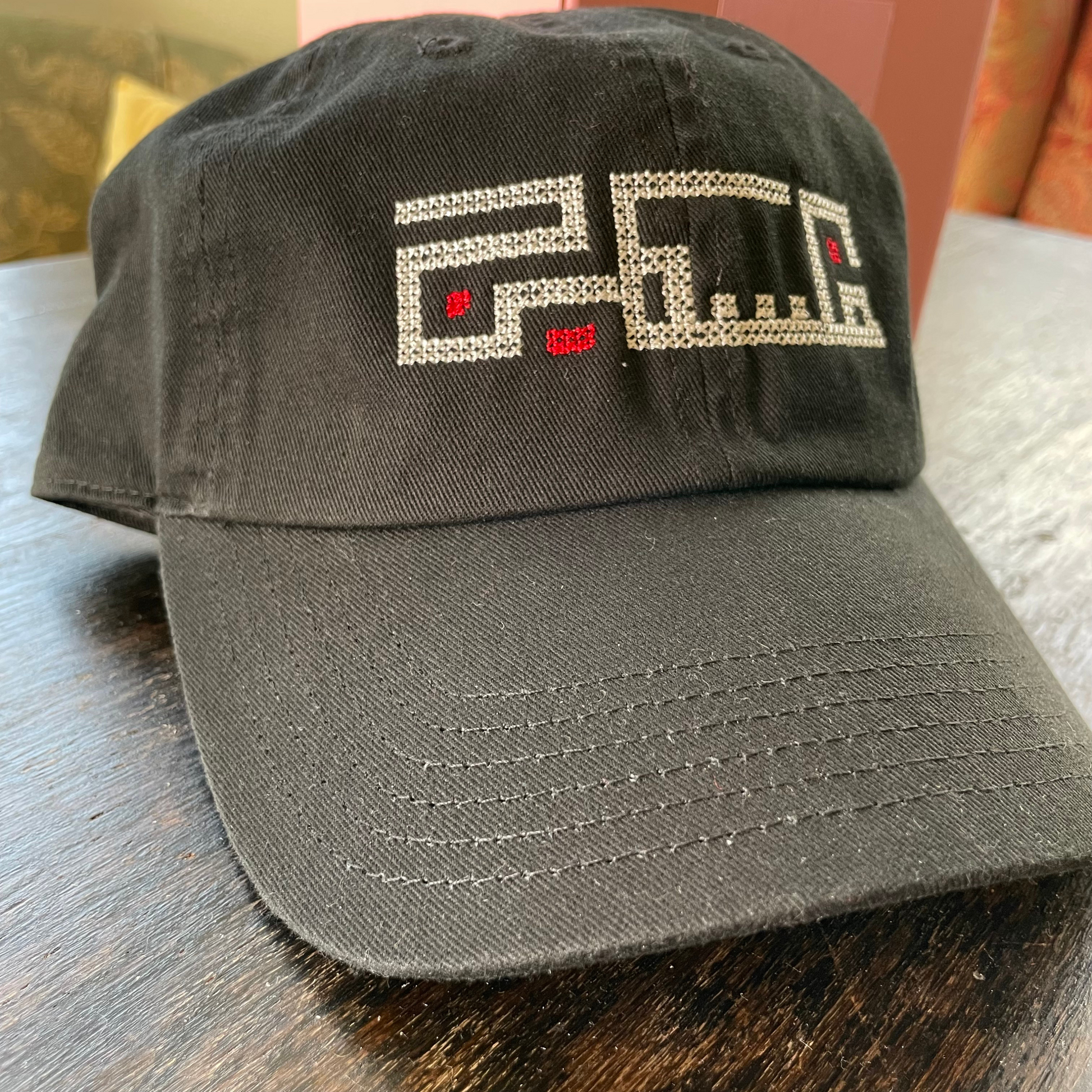 Palestinian embroidered black baseball hat, adjustable and made from 100% Cotton. This hat features an original embroidered design of the word "Palestine" using Arabic Kufic calligraphy. on a wooden table. view from the front