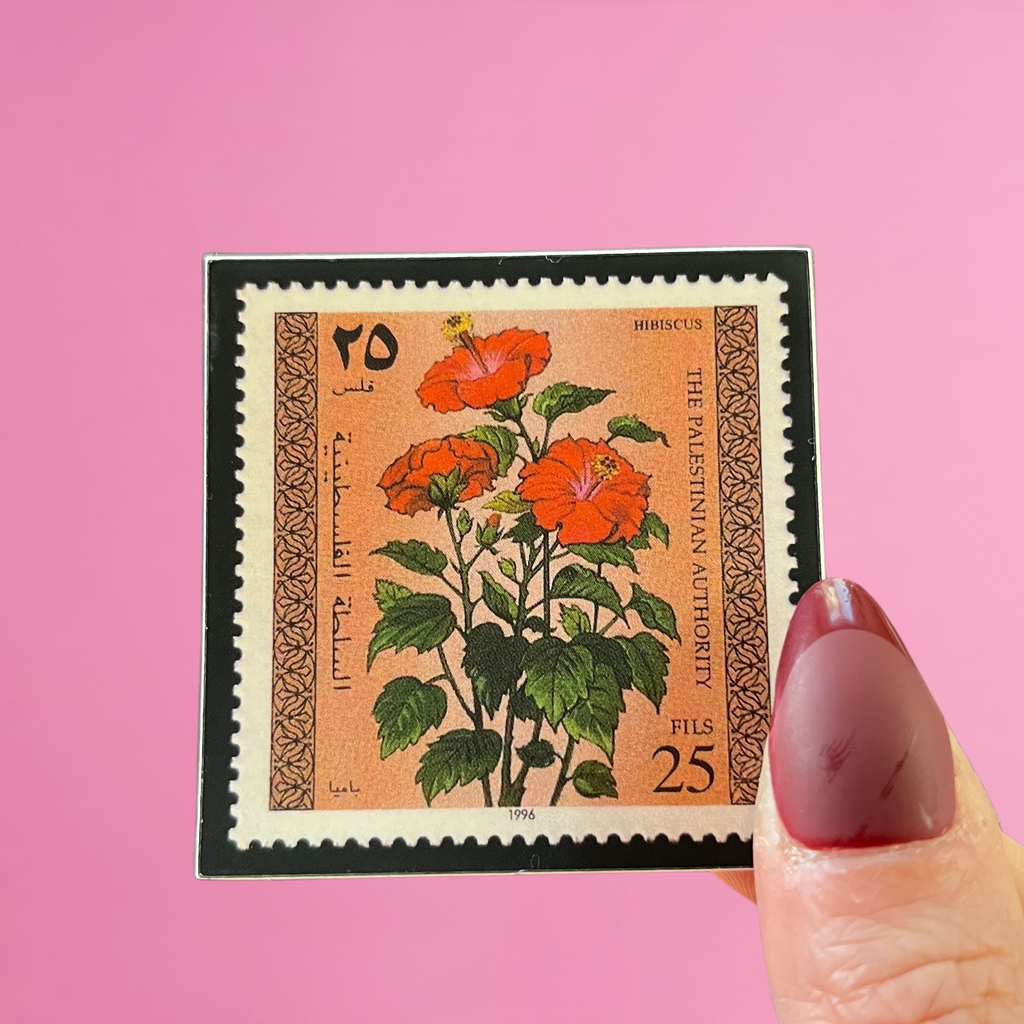 Front view of fingers holding a Palestine stamp with an image of a hibiscus flower. Against a pink background