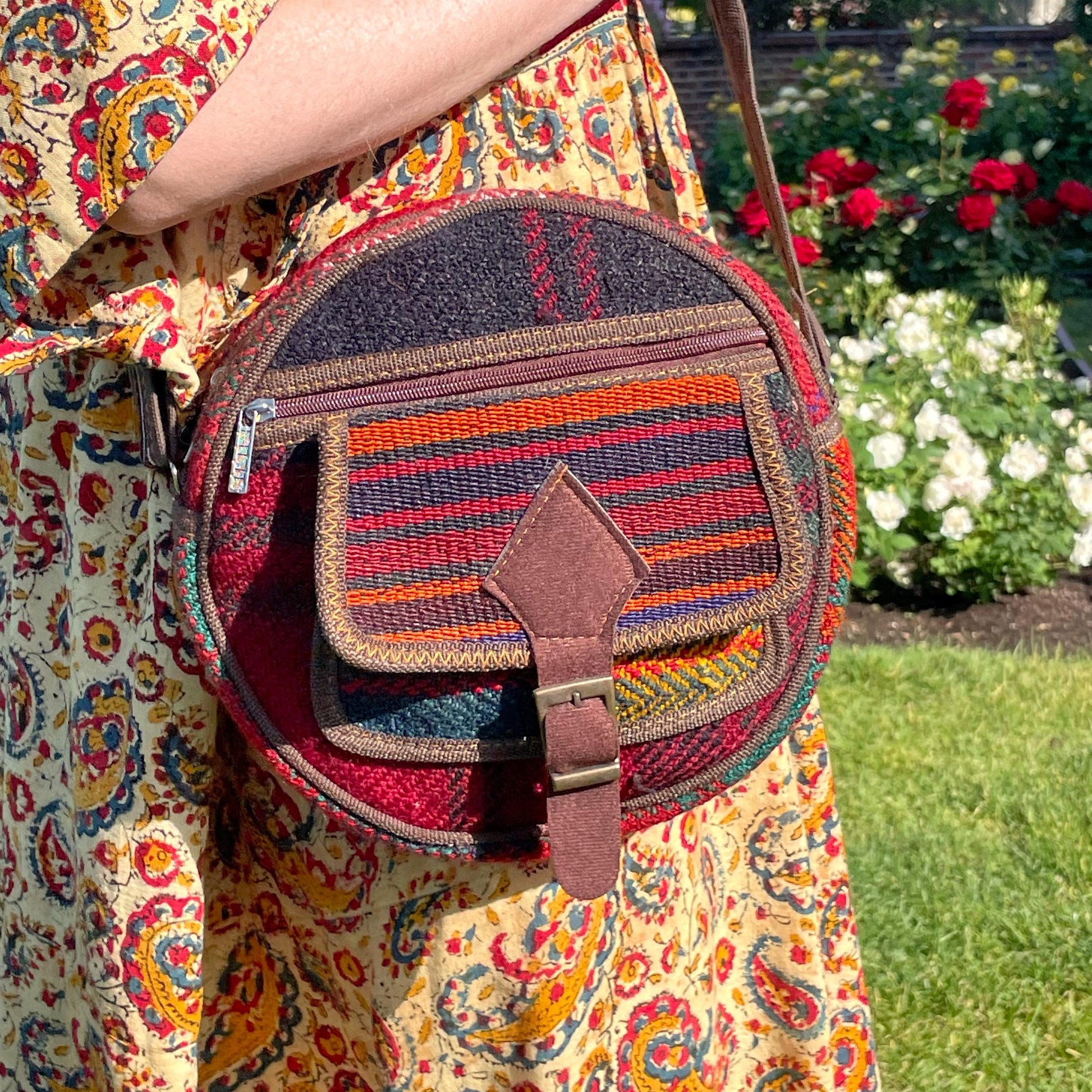 view from the front of a colorful circle bag modeled on a woman in a garden. the purse has handwoven sections of wool, trimmed with brown fabric. The bag has a long strap, front pockets, zippers, and a faux buckle over a snap closure